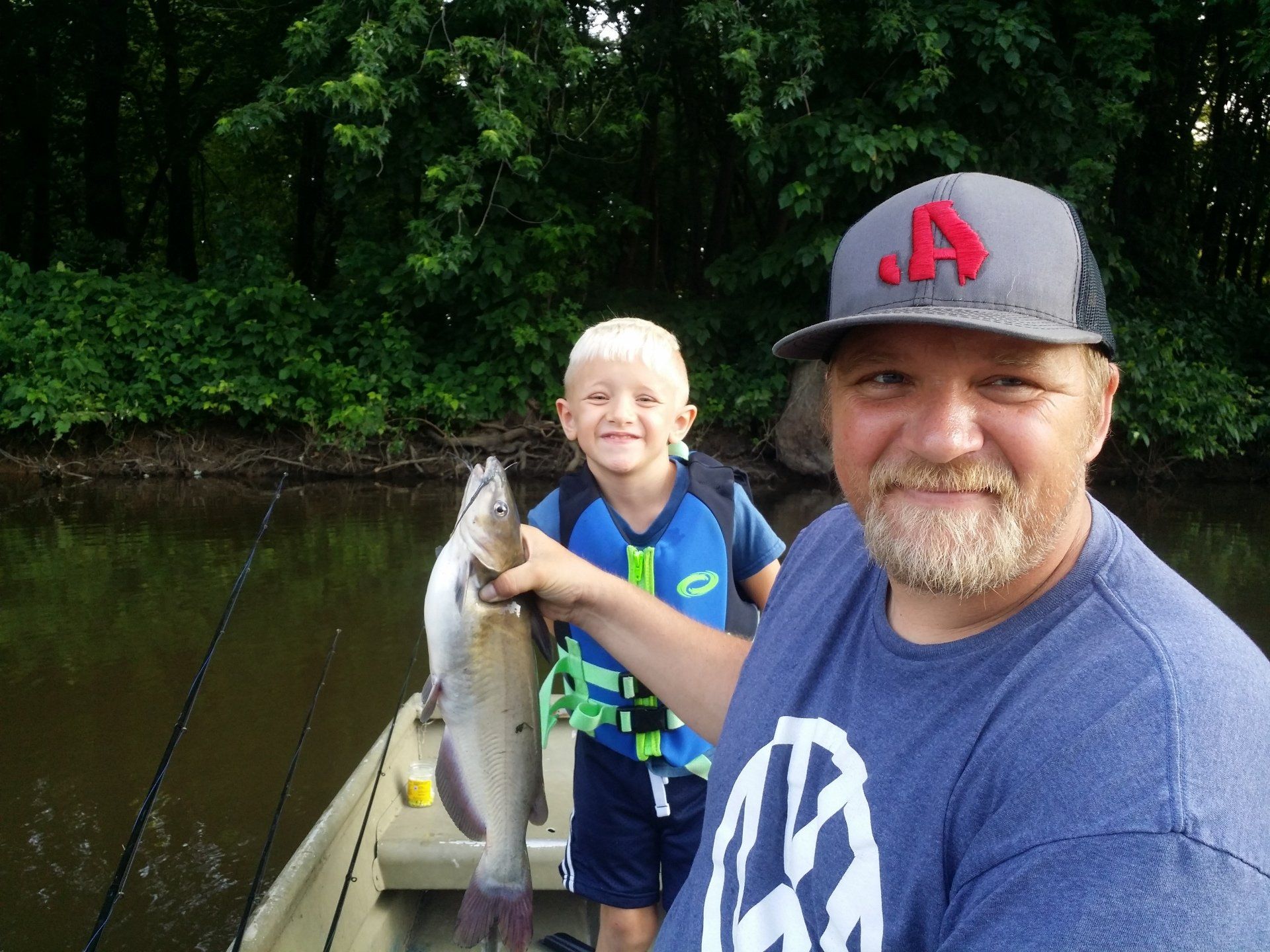 Man and child on a boat hold up a fish, smiling. Green trees and water in the background.