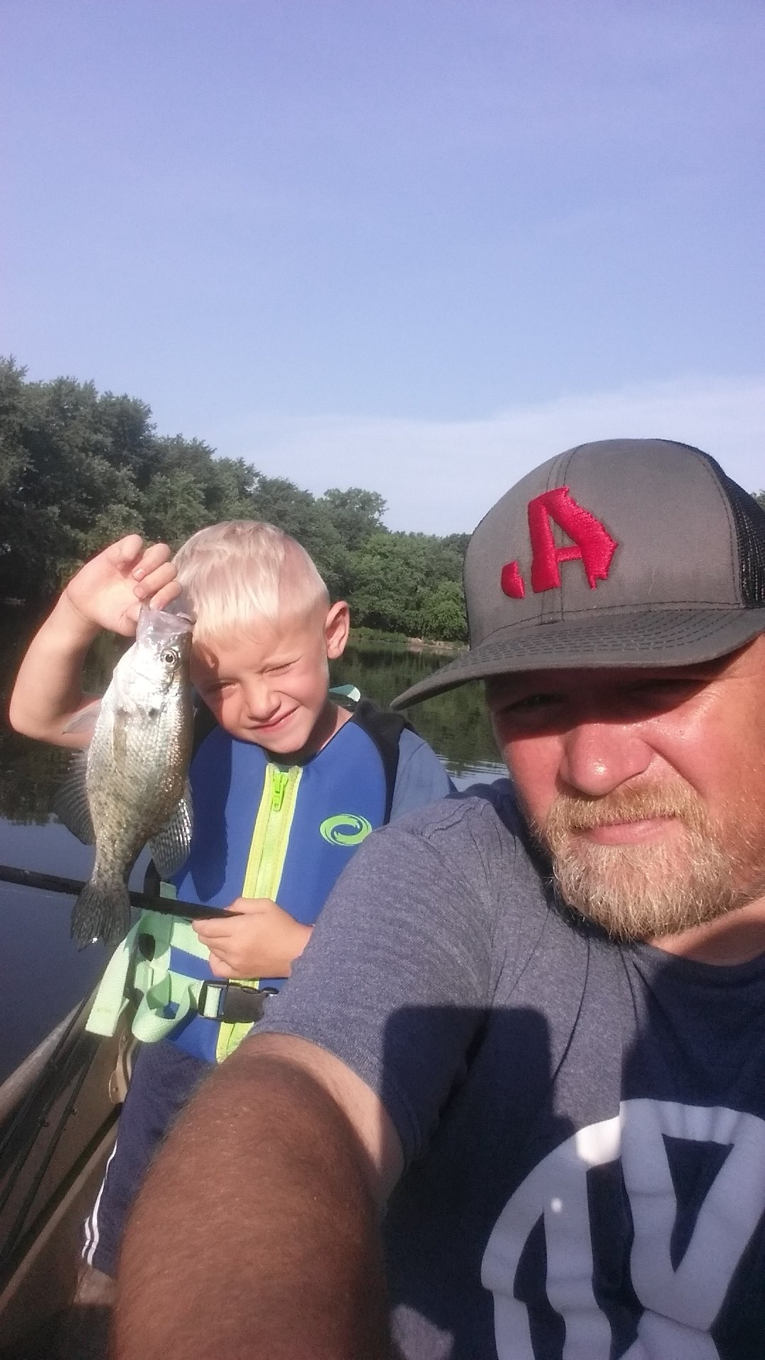 Boy holding up a fish, smiling on a boat with an adult. Trees and blue sky visible.
