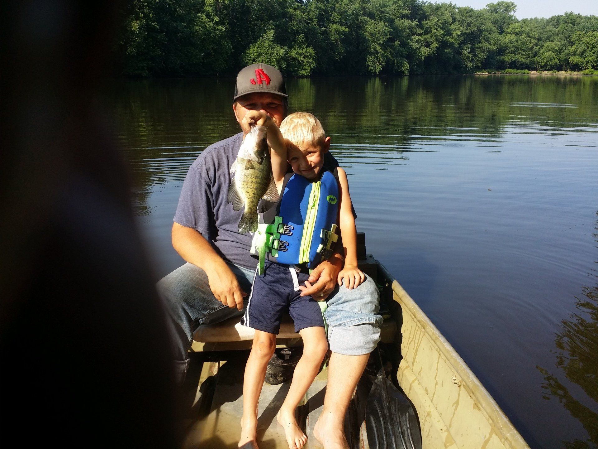 Man and child on a boat, holding up a fish. They are on a lake with trees in the background.