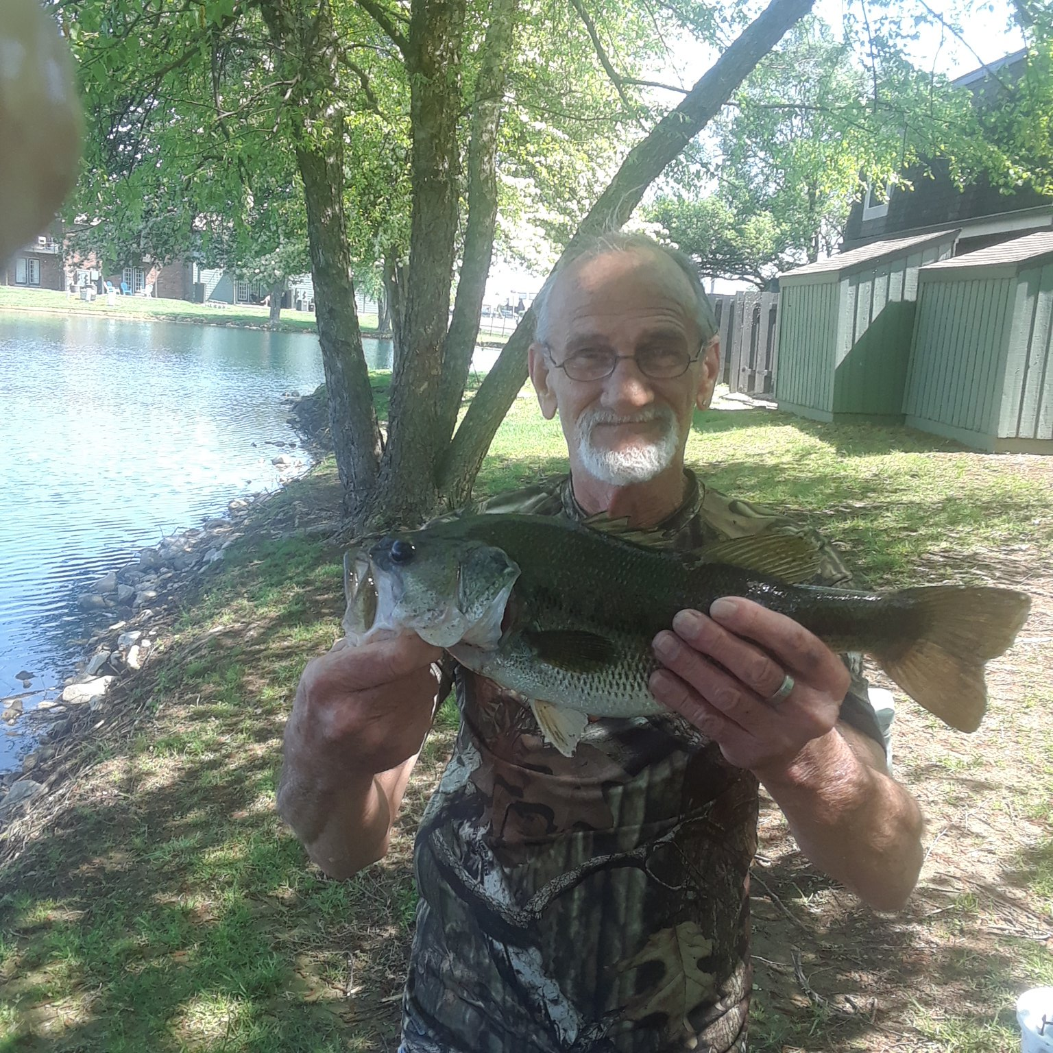Man holding a large bass fish near a pond. He wears camouflage and glasses, smiling.