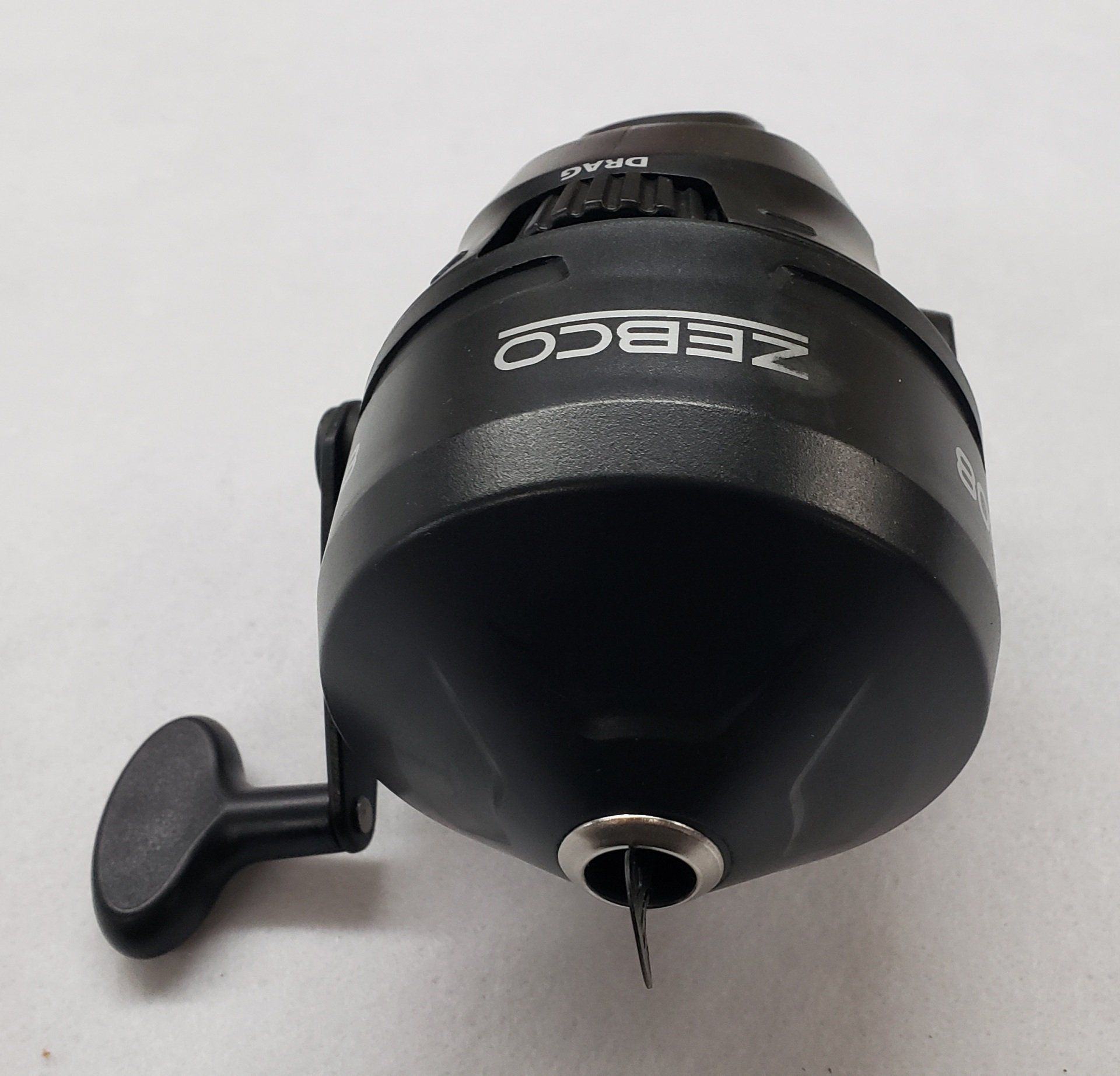 Black Zebco fishing reel with a handle on a white surface.