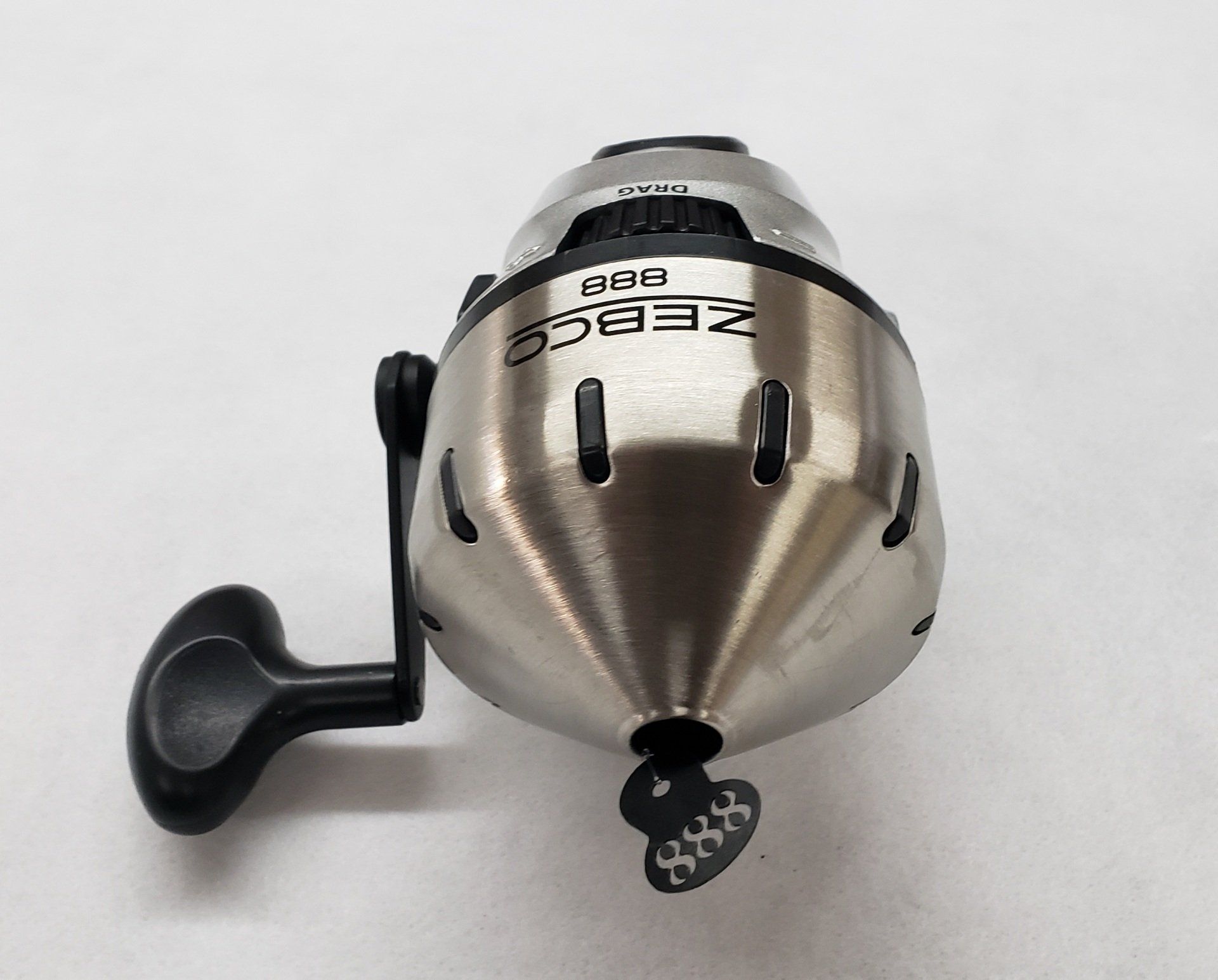 Silver Zebco Riba fishing reel with black handle, sitting on a white surface.