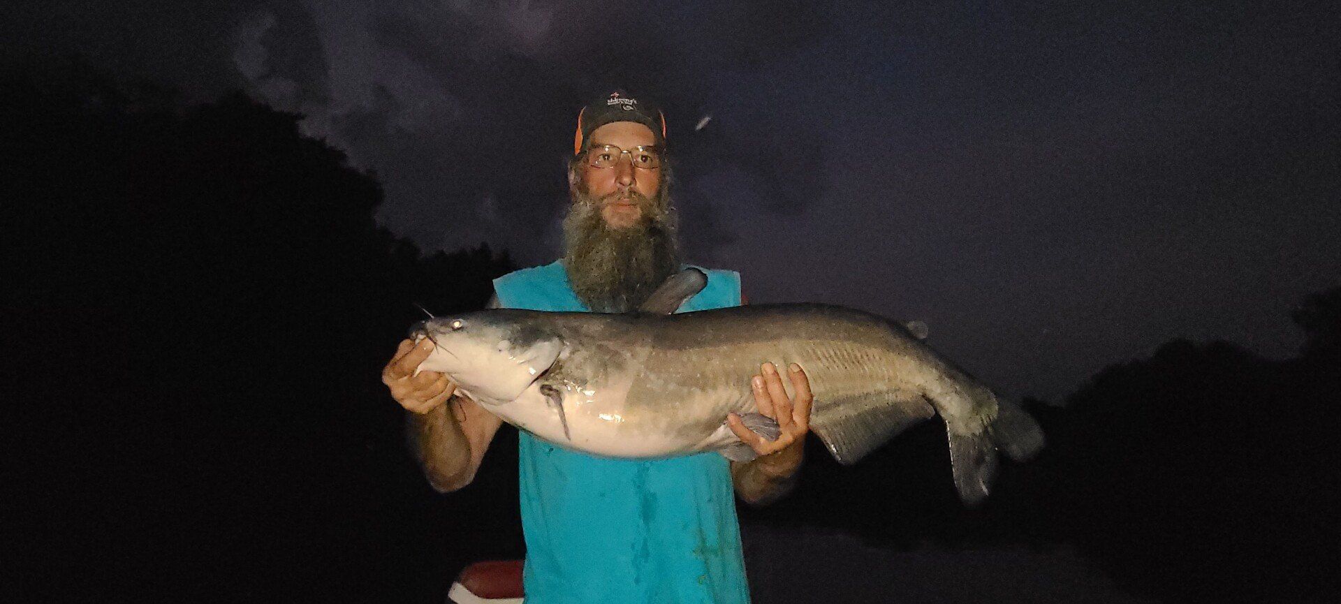 Man holding a large catfish at night, with dark sky and blue shirt.