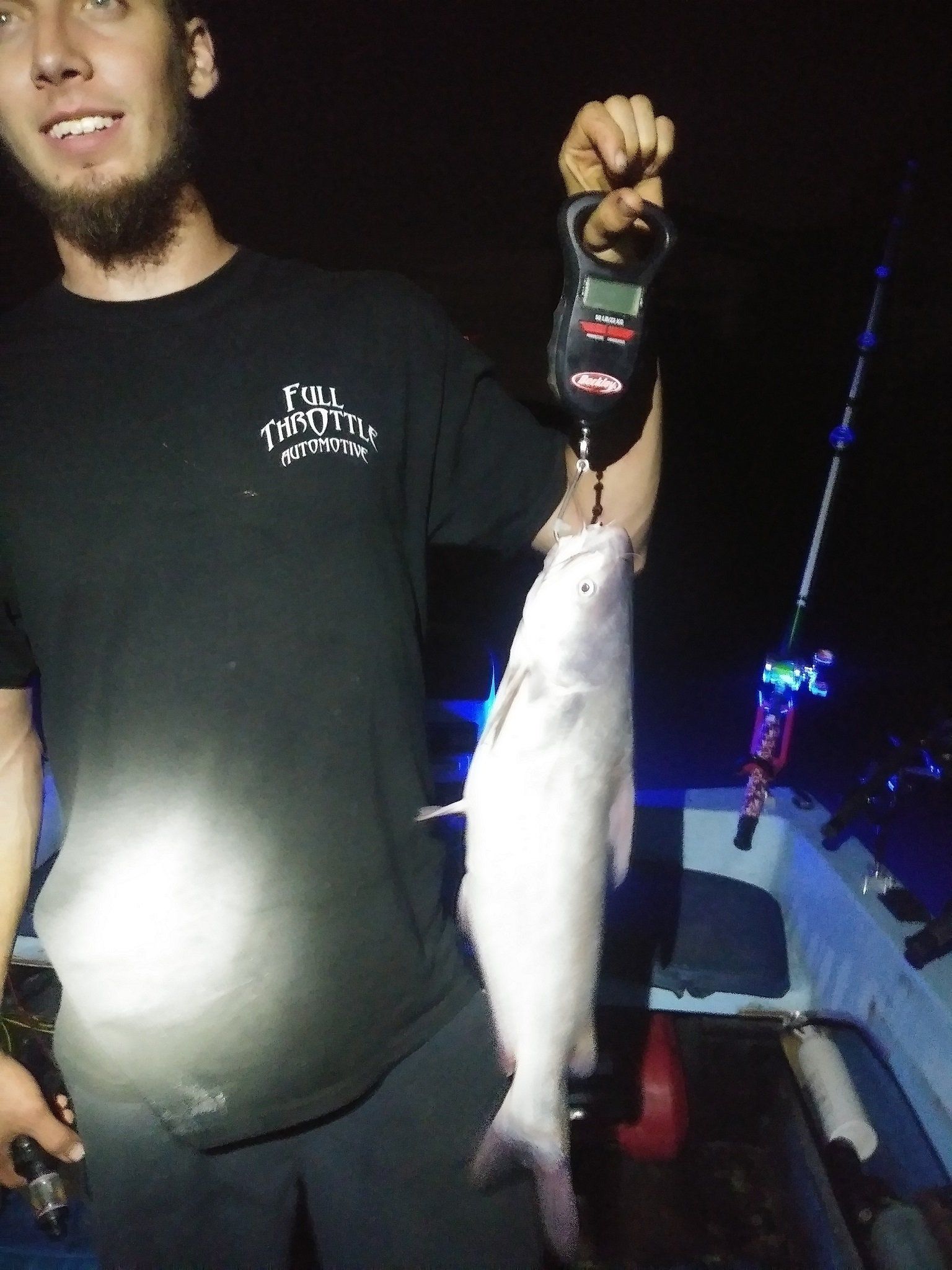 Man holding a fish on a scale at night; on a boat, a fishing rod visible.