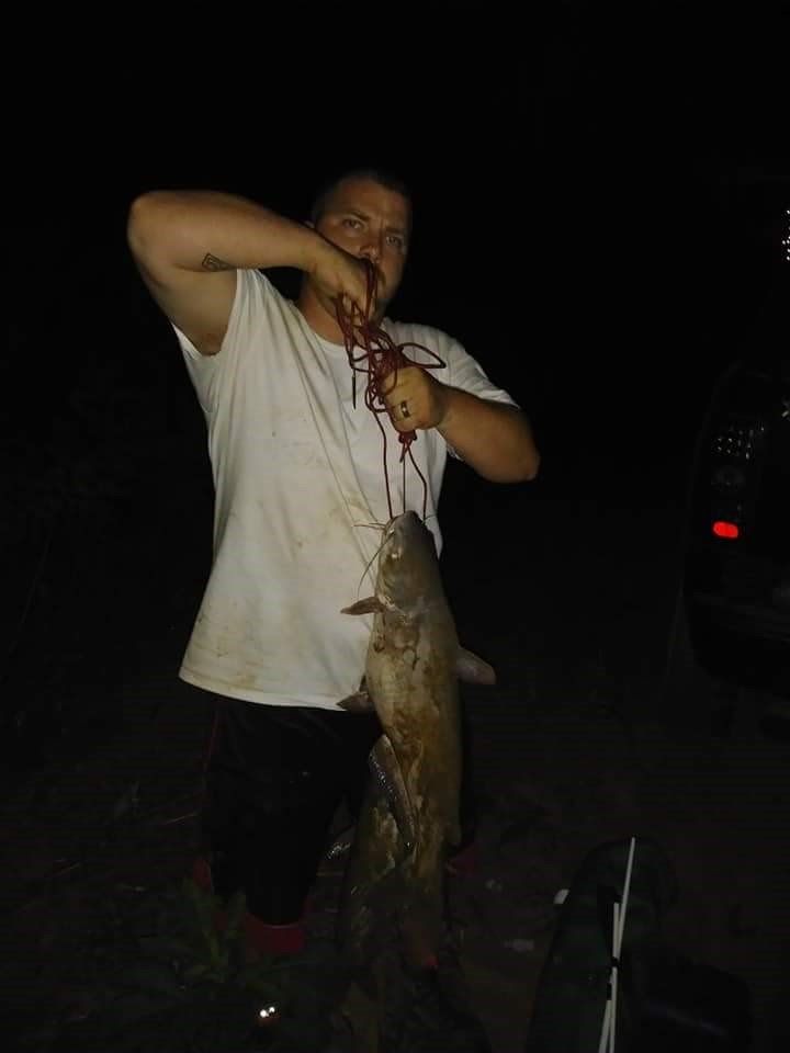 Man holding a large fish by rope, posing outdoors at night. Dark, shadowy setting.