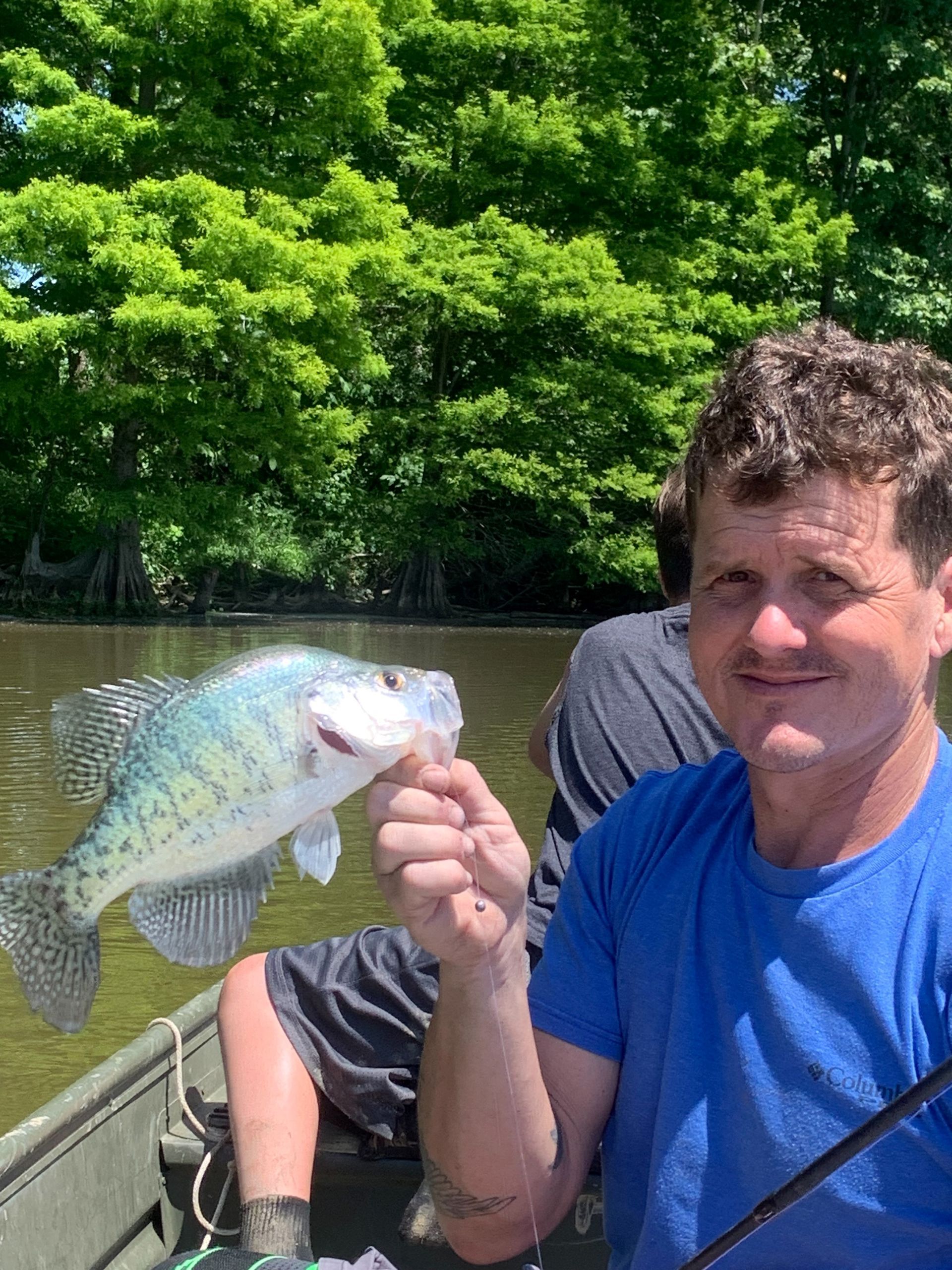 Man in blue shirt holding a spotted fish on a boat, with trees in the background.