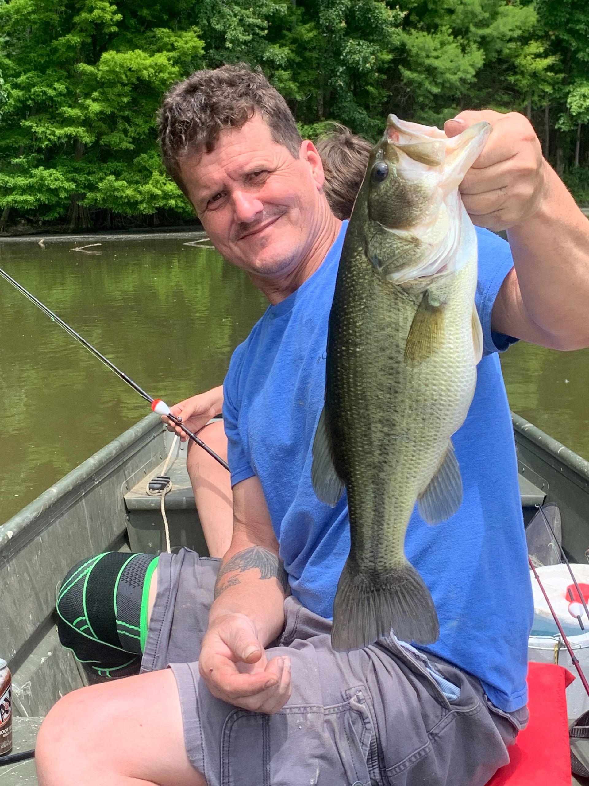 Man in a boat holds up a largemouth bass he caught. Green trees and water in the background.