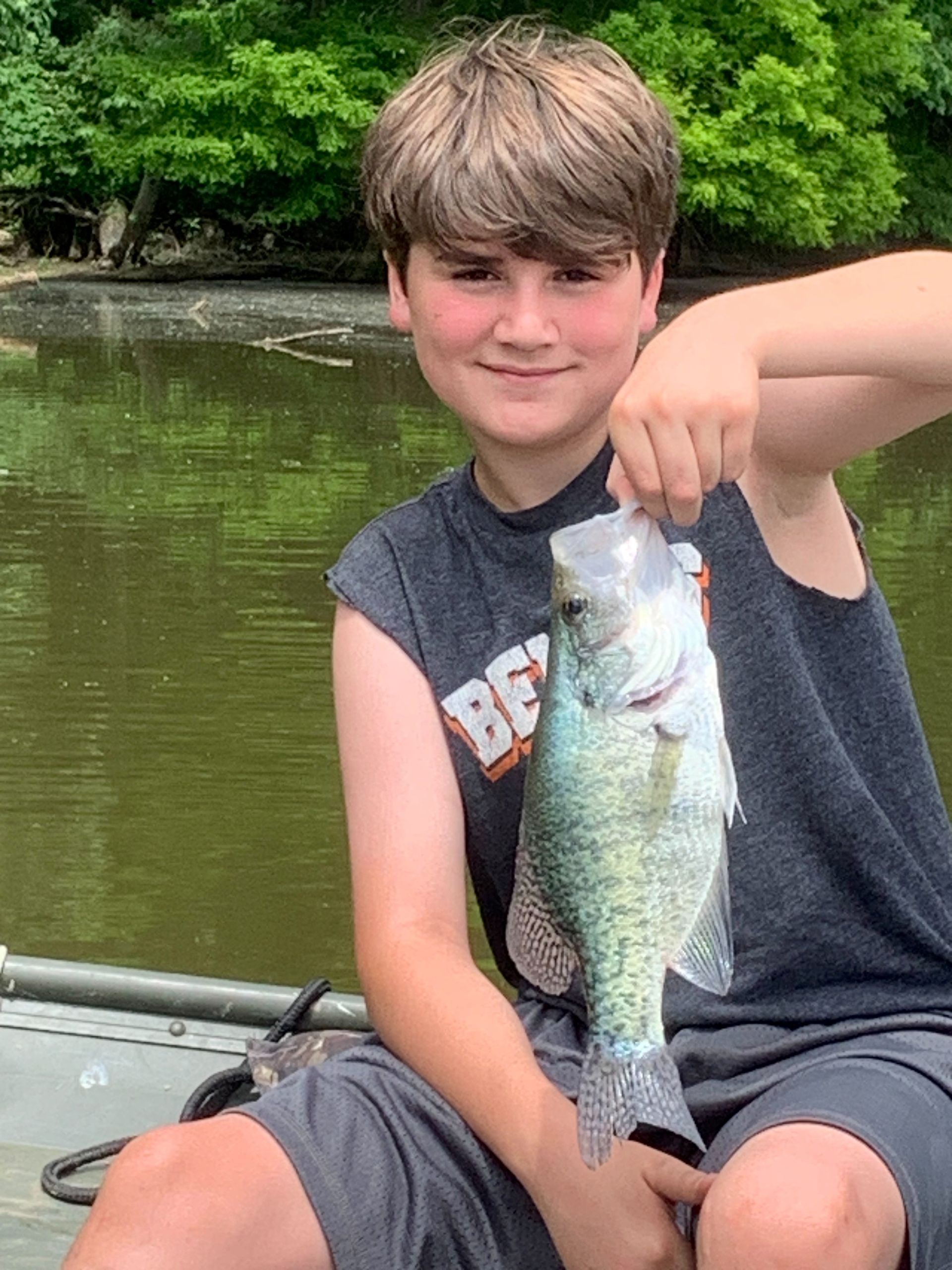 Boy in sleeveless shirt holding a crappie fish, on a boat, green water in background.