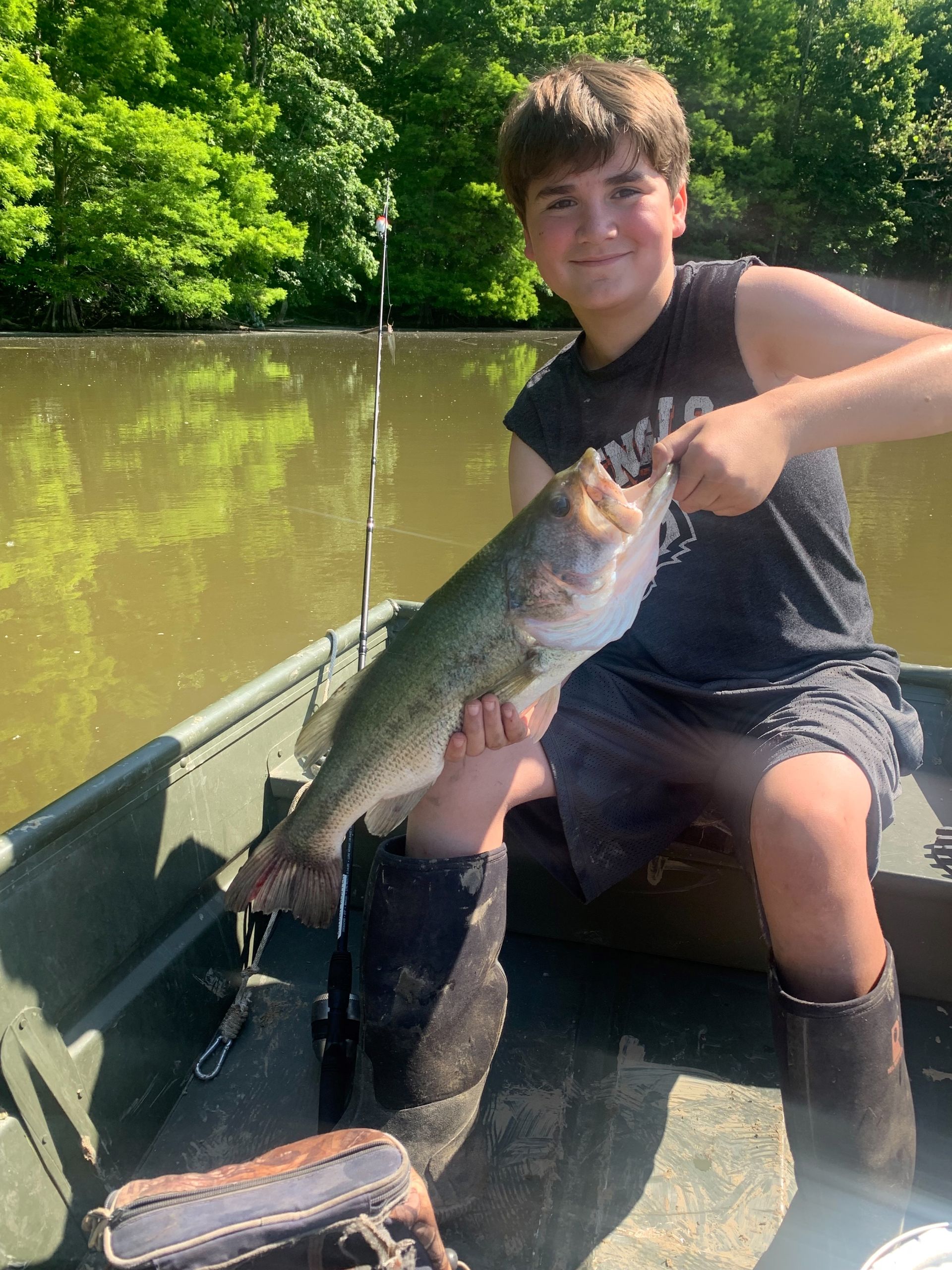 Boy in boat holding a large fish, smiling. Green trees and water background.