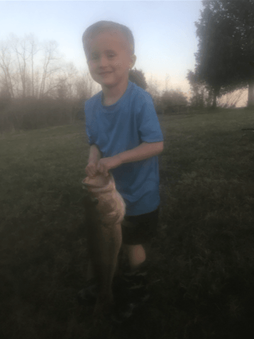 Boy smiling, holding a large fish, standing on grass. Dusk setting, trees in background.