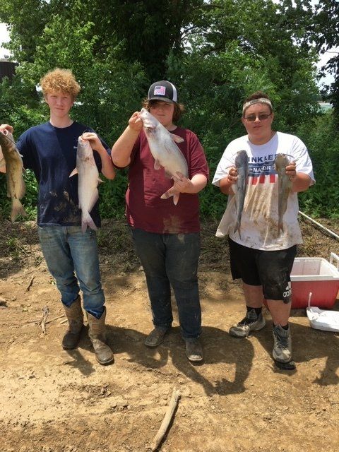 Three people holding fish, standing outside near a body of water.