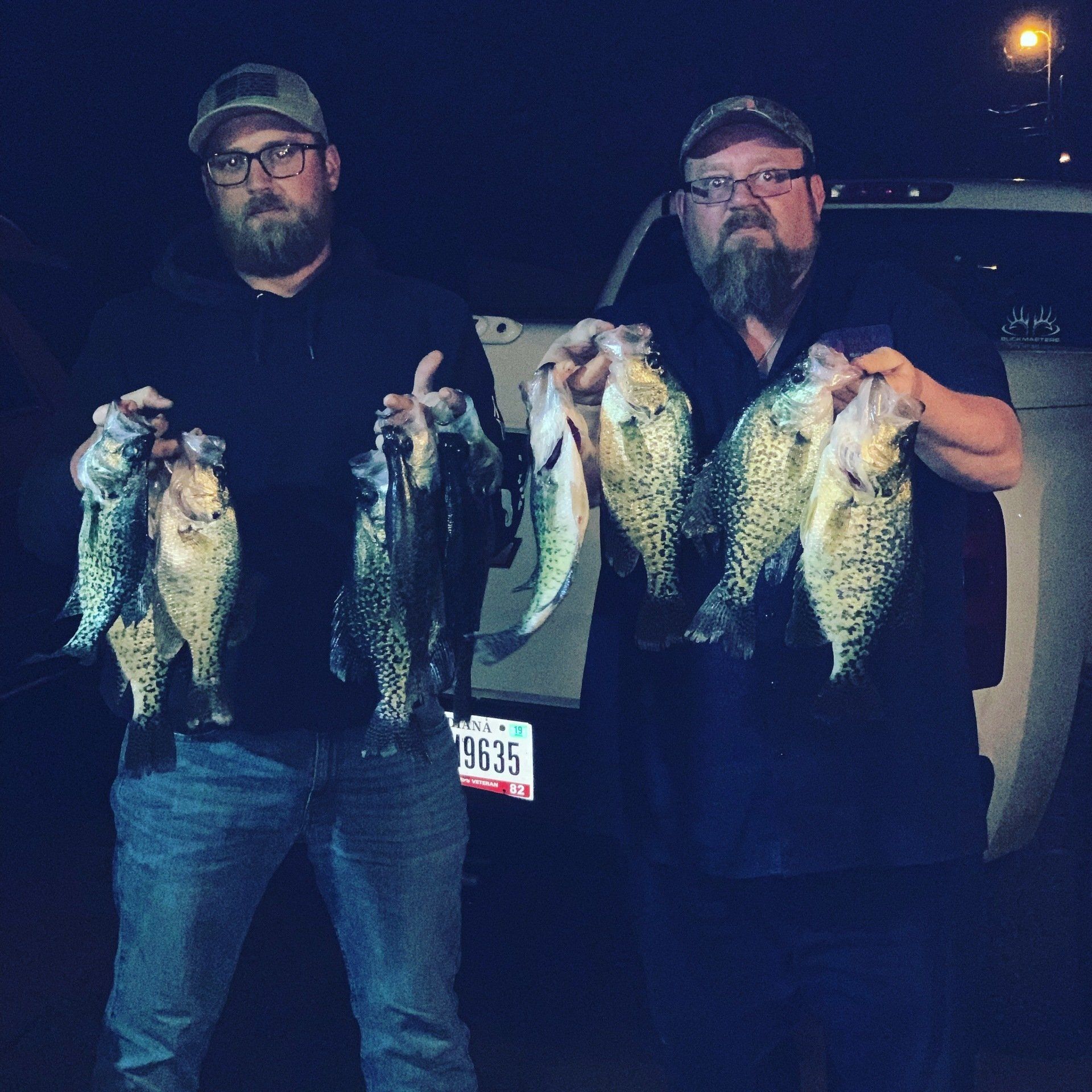 Two men holding up a large catch of speckled fish at night. They are outside next to a car.