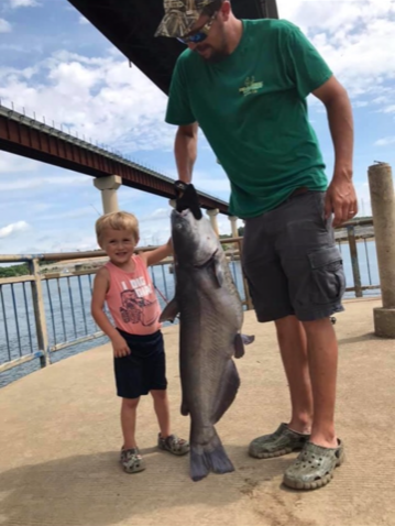 Man and child holding a large catfish on a pier. Blue sky, water visible.