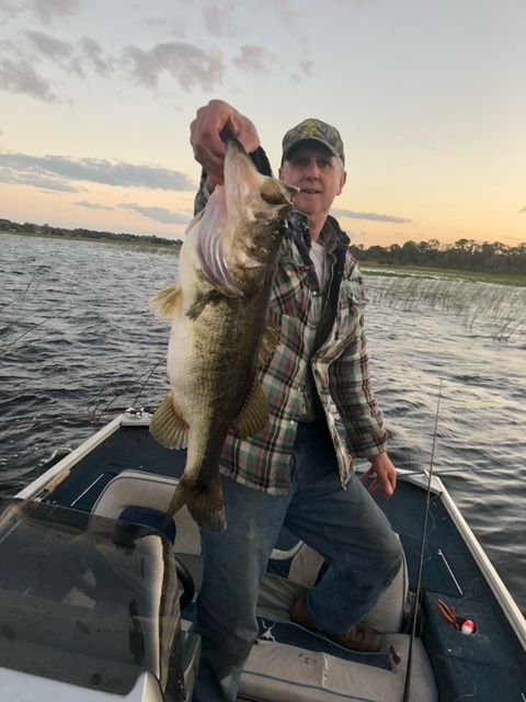 Man in boat holds up a large bass fish. Setting is a lake at sunset.