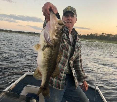 Man on boat holding up a large fish, presumably a bass, with lake and sky visible.