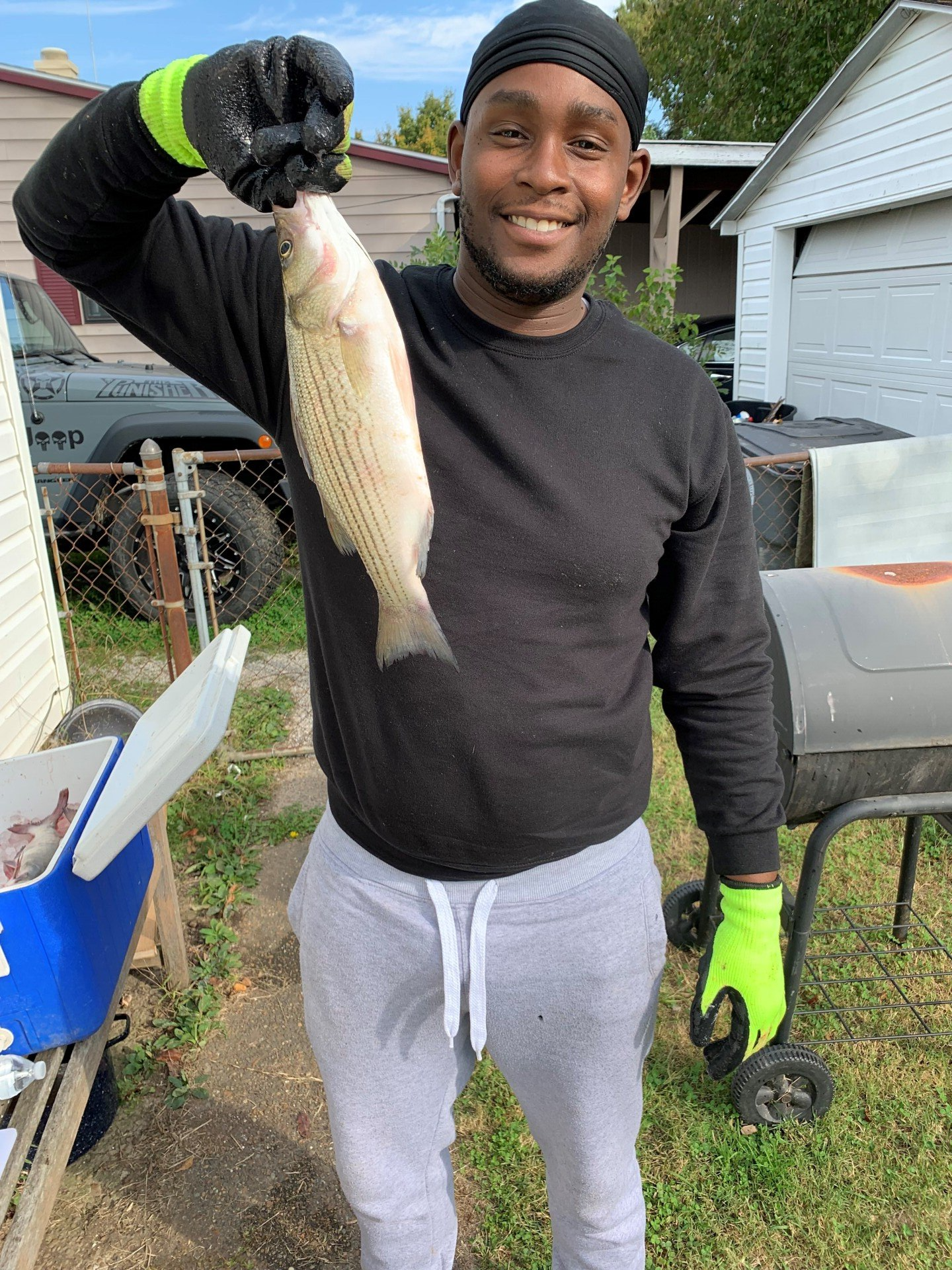 Man smiling, holding a fish, wearing black long-sleeve, gray sweats, and black cap outdoors.