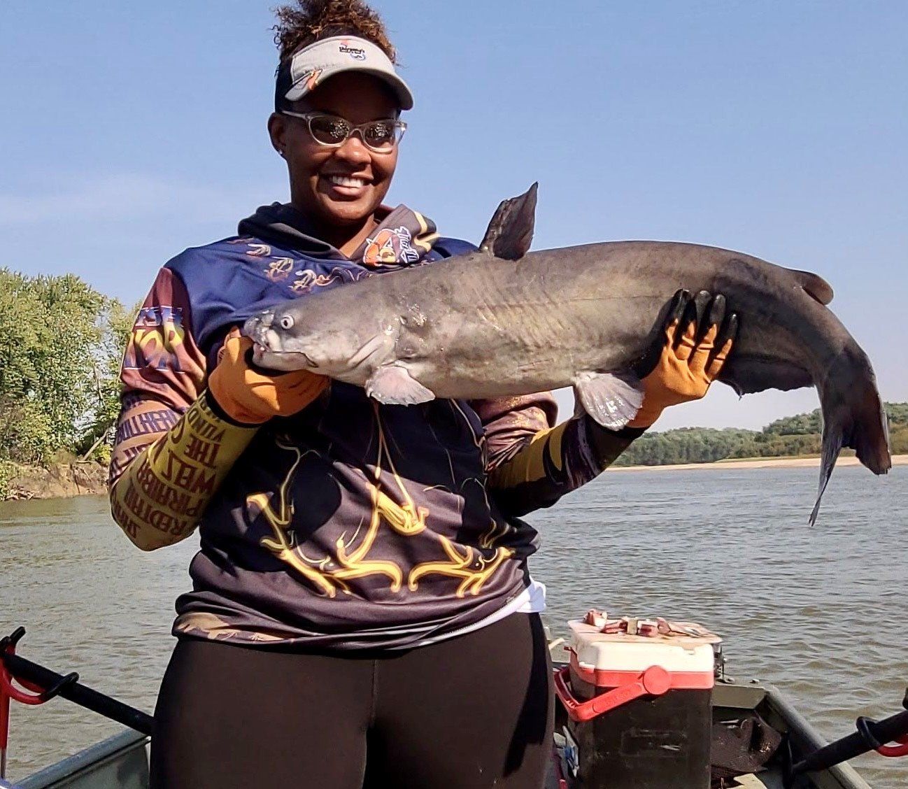 Woman on a boat holding a large catfish, smiling. Outdoors, sunny, water visible.