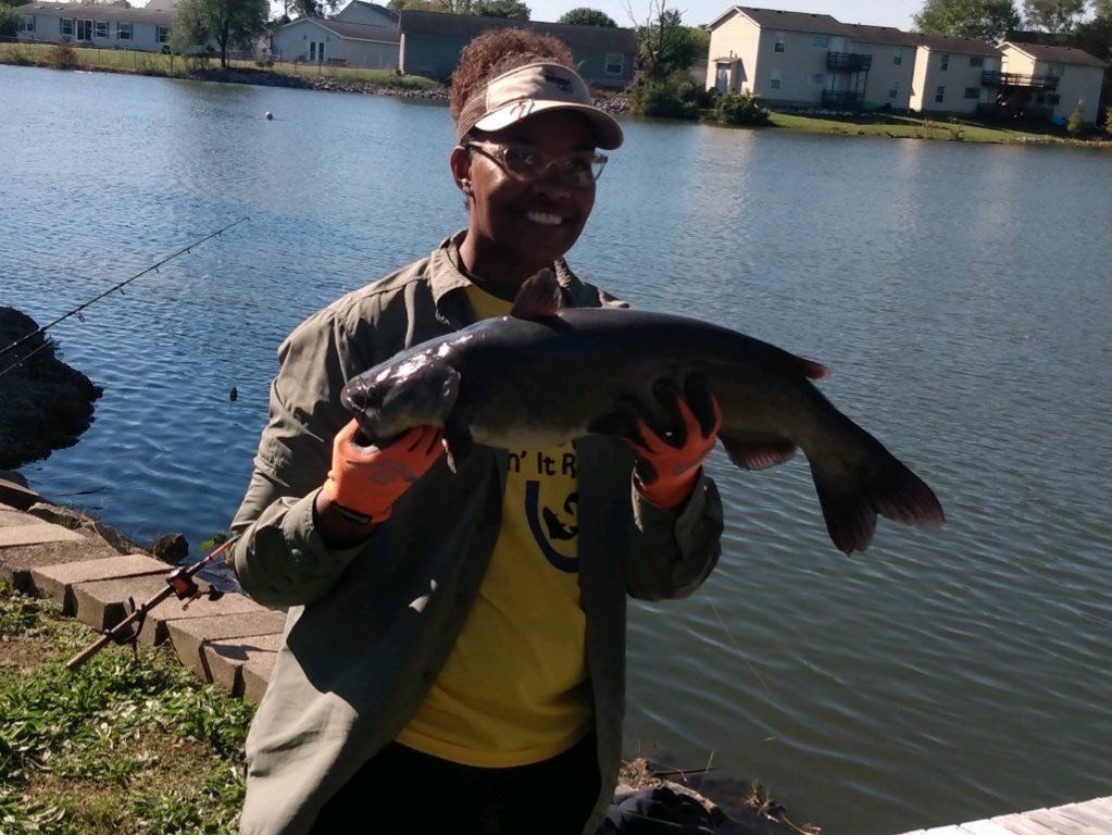 Woman holding a large fish near a lake; she is smiling, wearing a visor and gloves.