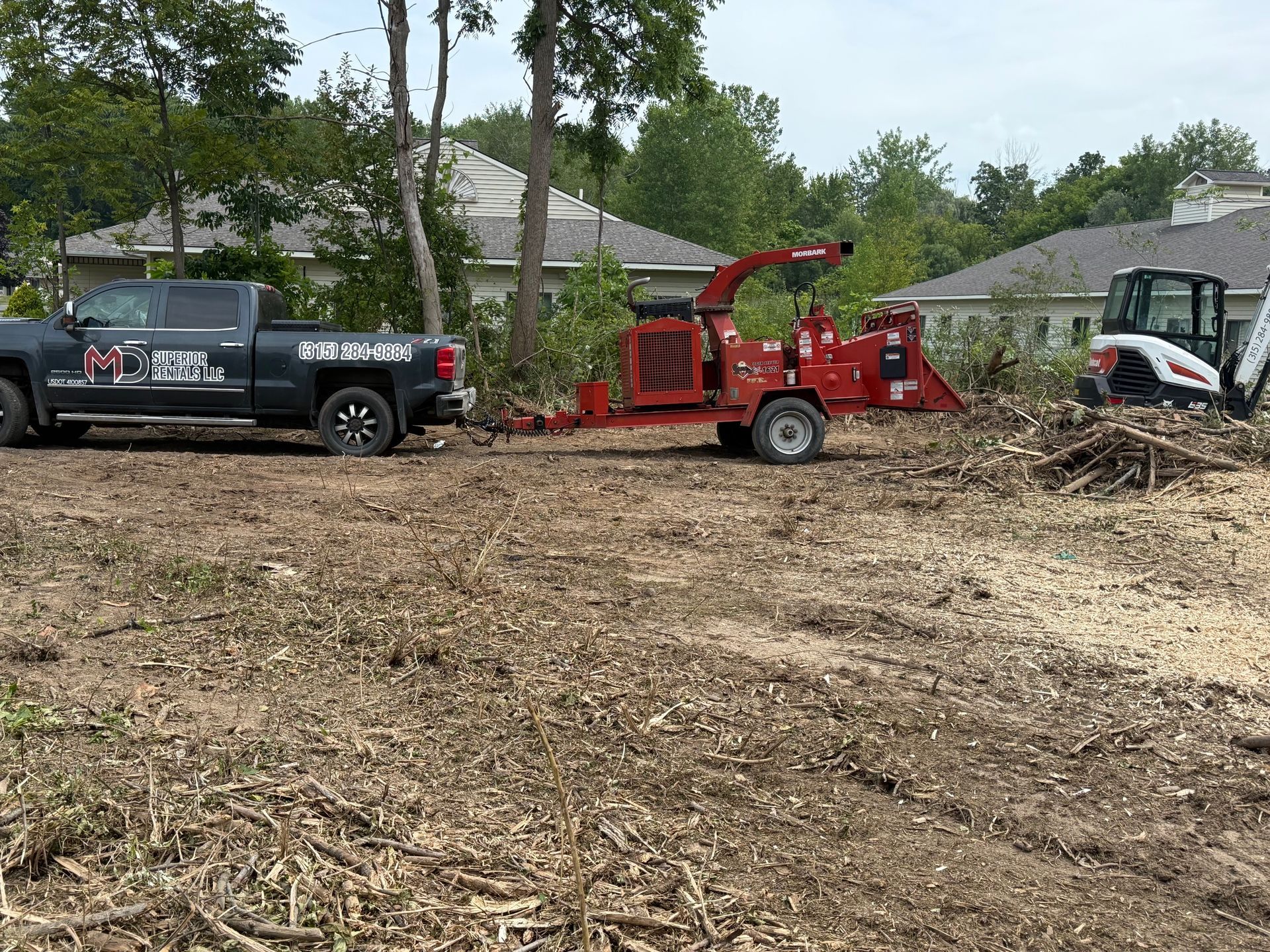 A black truck towing a red wood chipper next to a Bobcat, all on a cleared lot with debris, possibly tree removal.