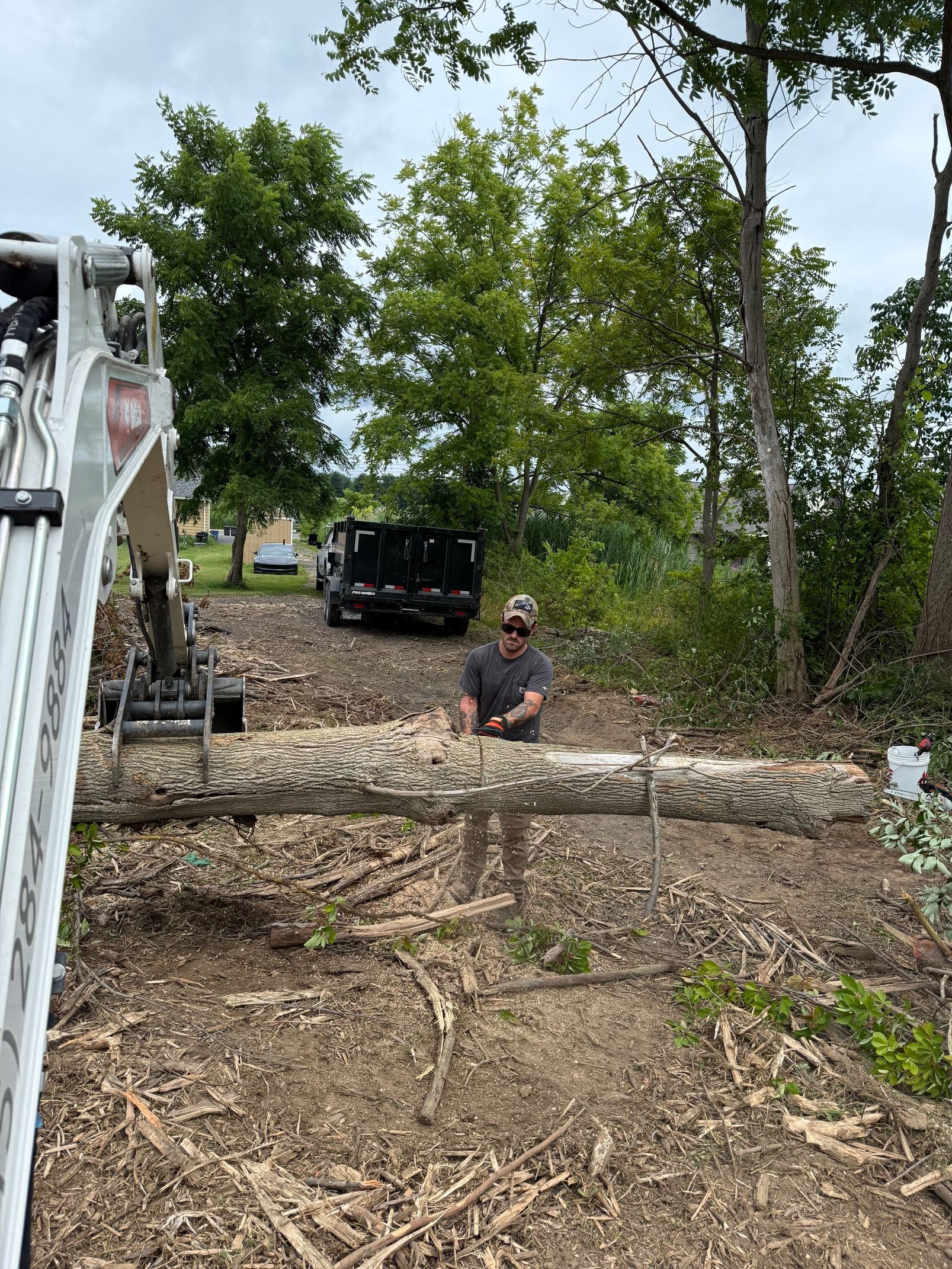 A person with a hat cuts logs outdoors with a chainsaw; an excavator arm holds logs, debris is on the ground, and a truck sits nearby.