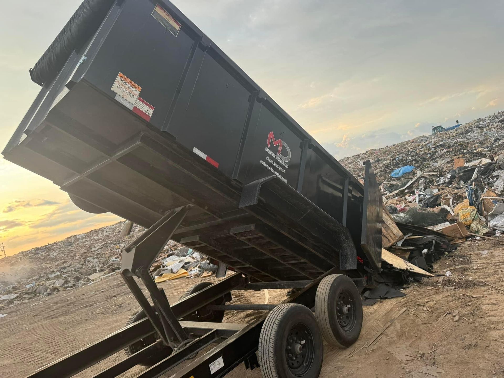 Black dump trailer unloading waste at a landfill, against a cloudy sky.