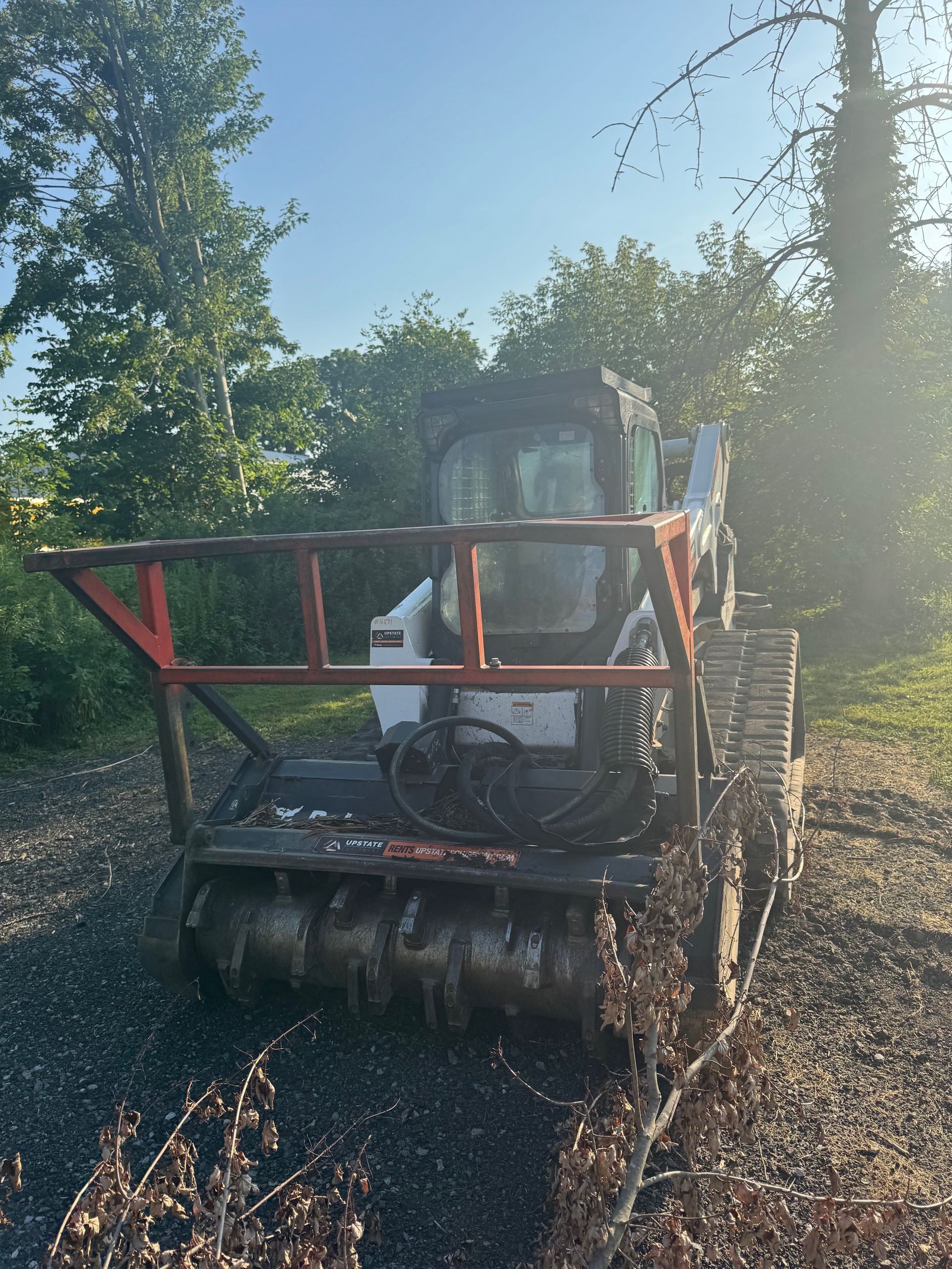 Bobcat with a brush cutter attachment, clearing a gravel path outdoors on a sunny day.