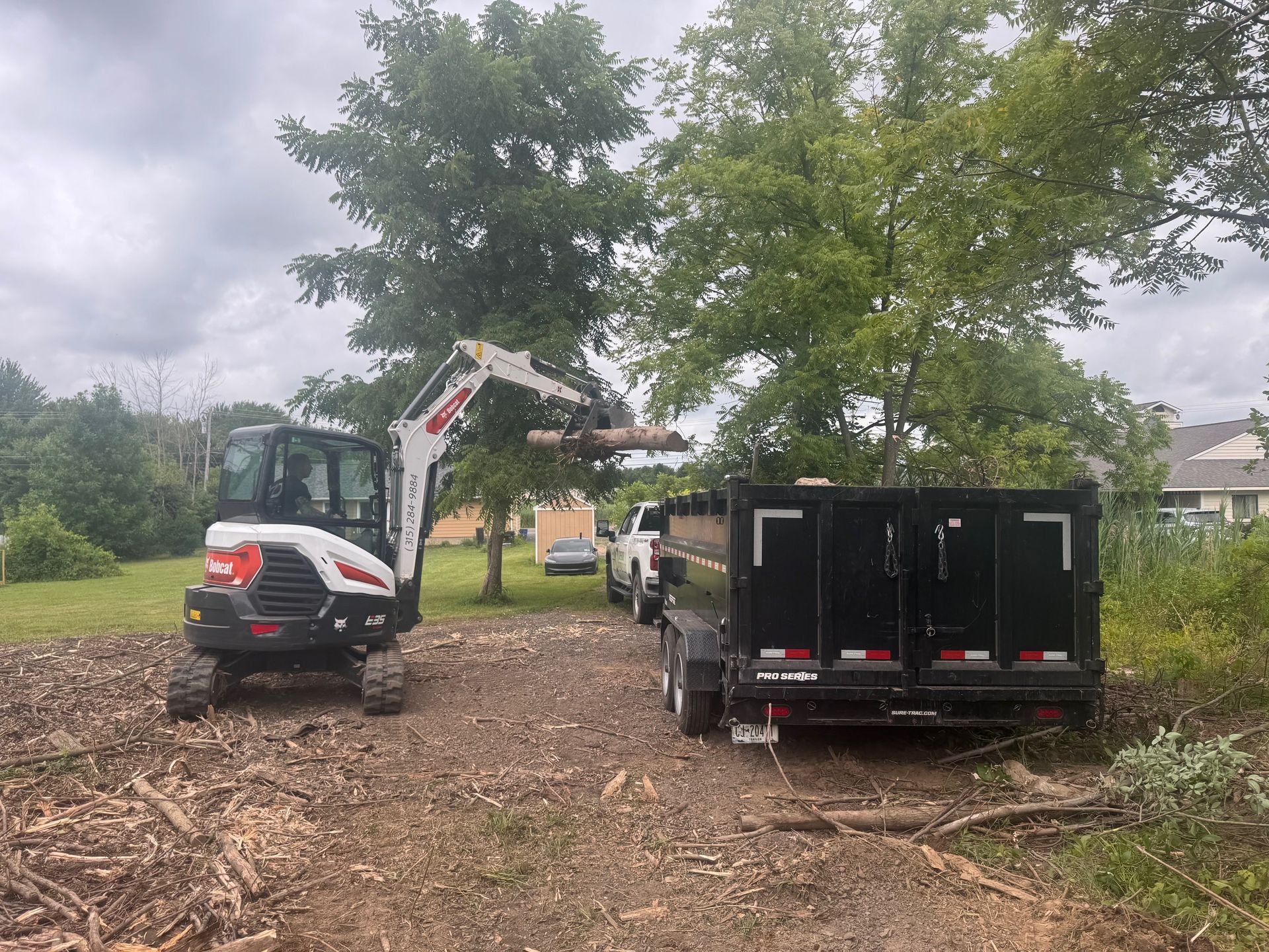 A Bobcat excavator loading tree branches into a black trailer on a wooded property.