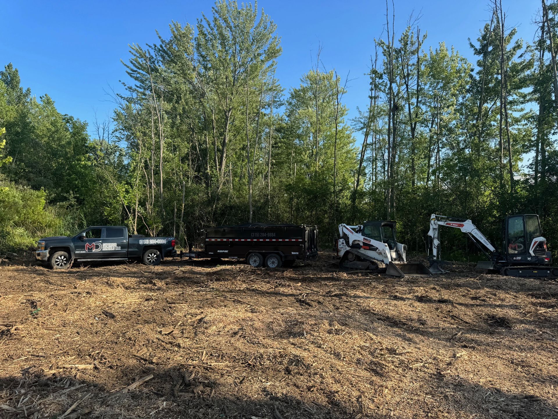 A work site with a truck, trailer, skid steer, and excavator in front of a wooded area. The ground is covered in wood chips.