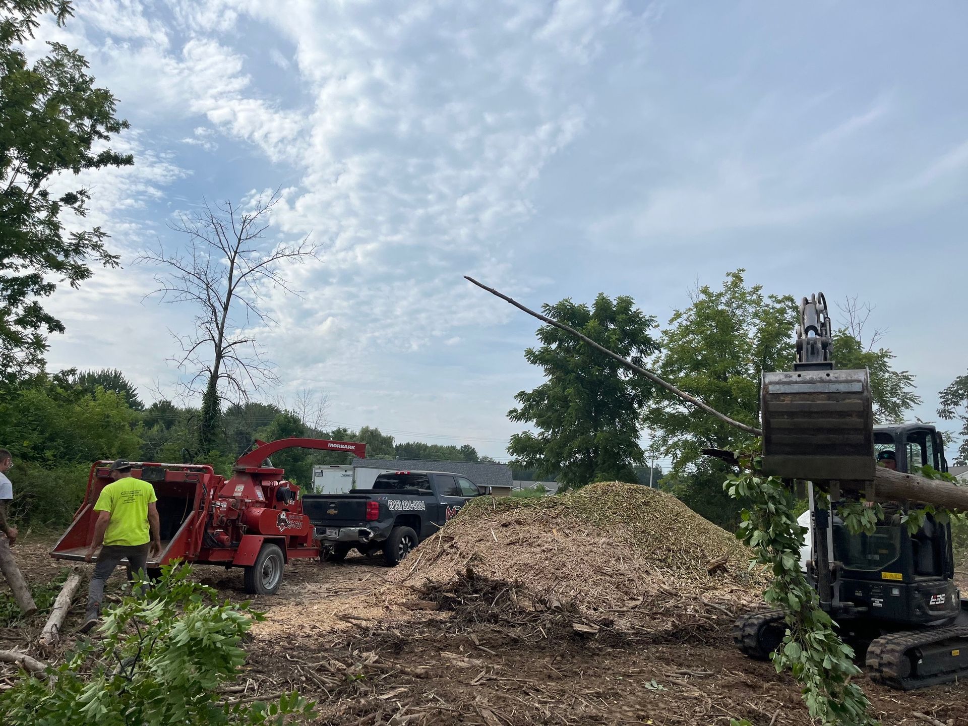 A tree service clearing a wooded area. A wood chipper, truck, and excavator are used to remove tree branches and create a large pile of wood chips.
