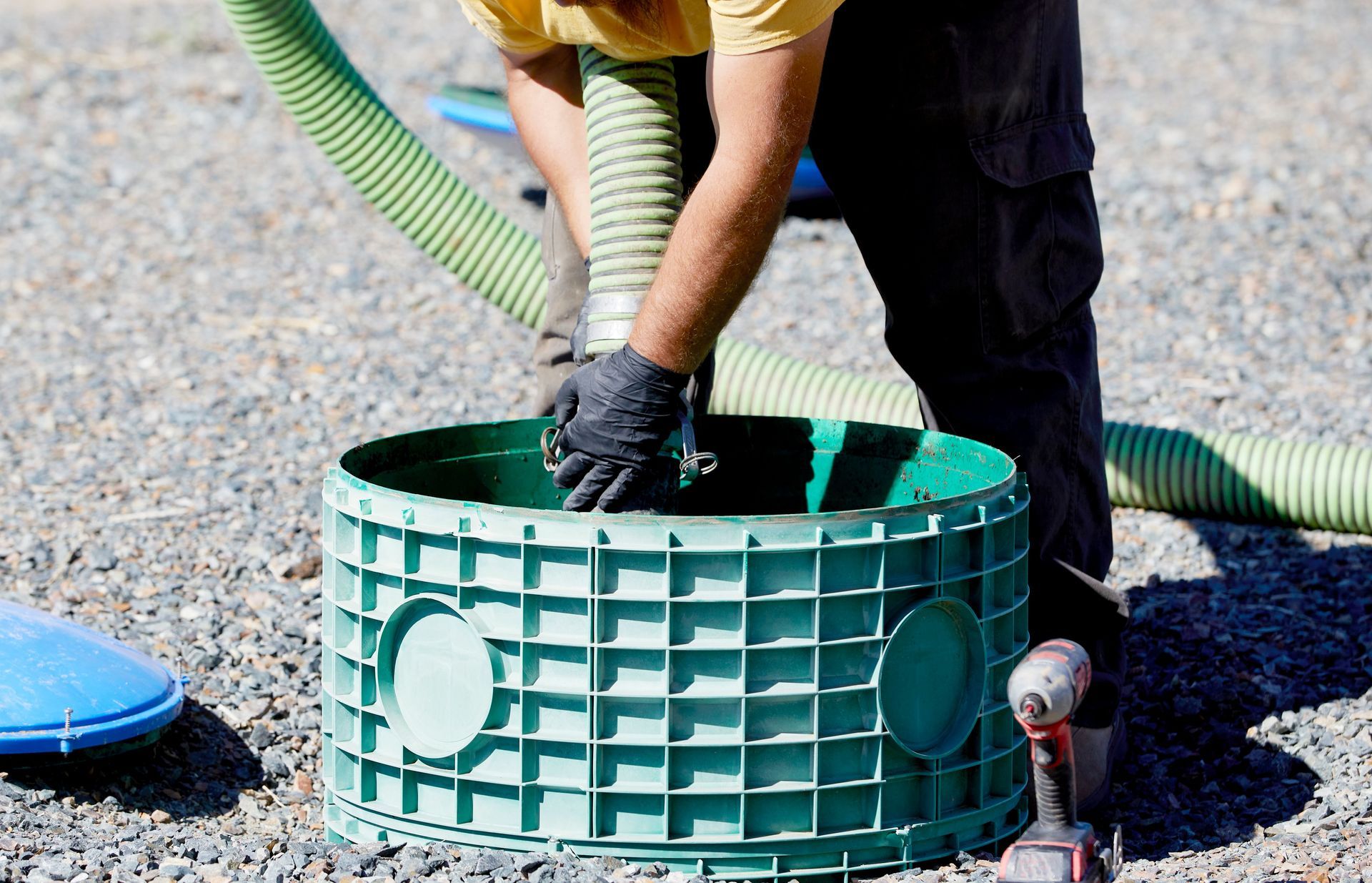 Person wearing gloves installs a green plastic drainage chamber on gravel outdoors