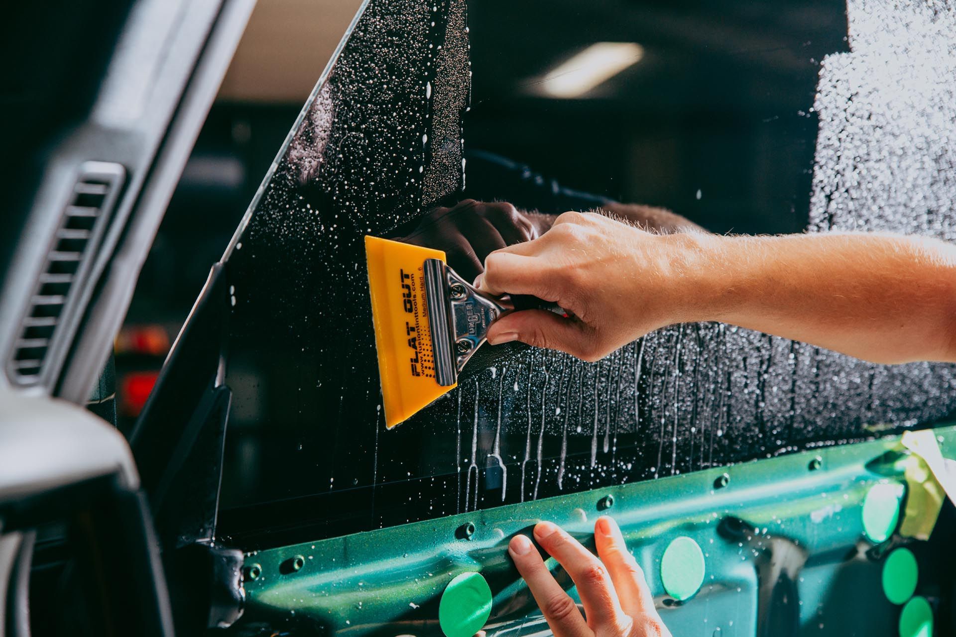 A person uses a yellow squeegee to smooth window tint film onto a car window covered in soapy water.