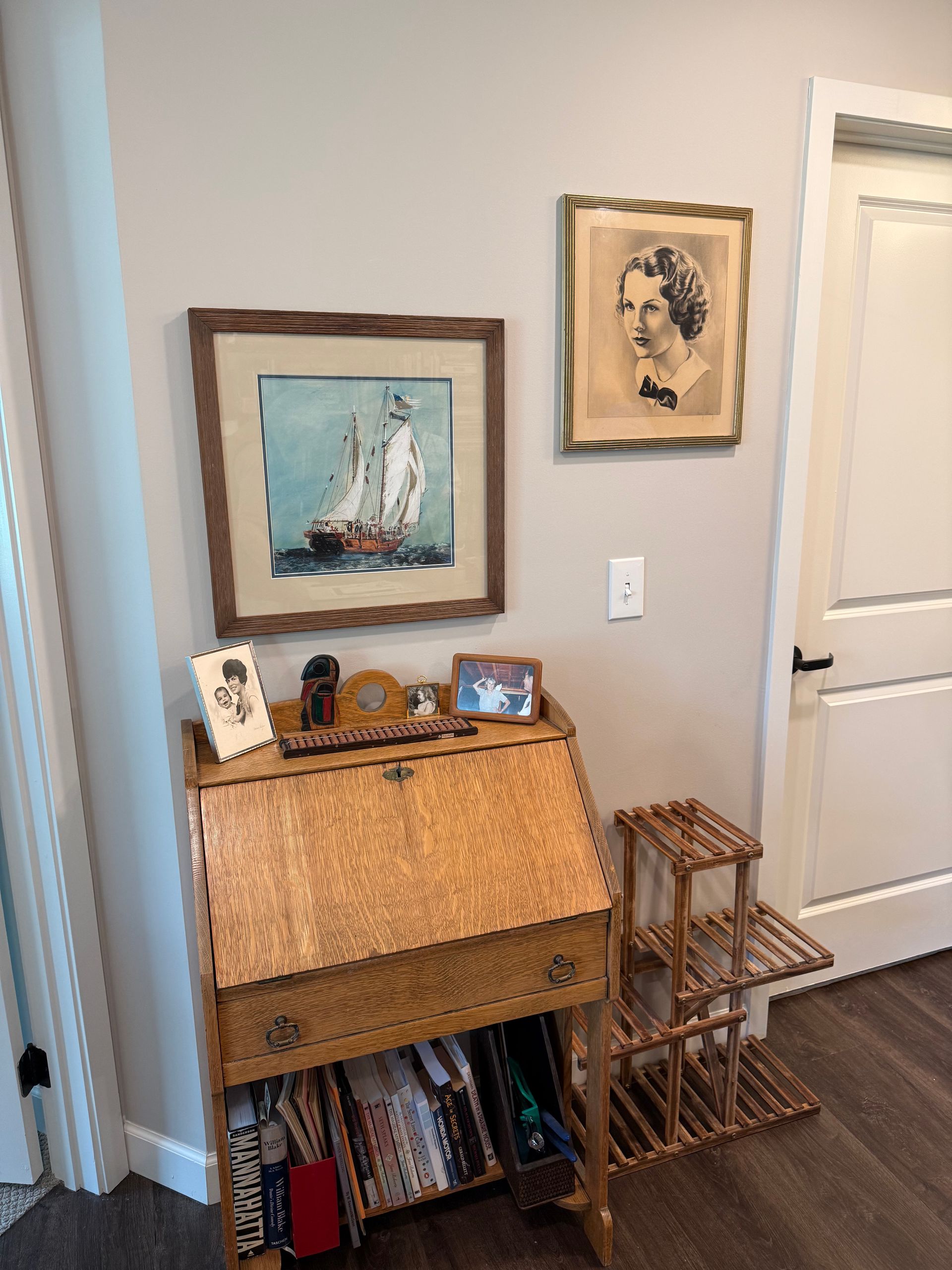 Wooden desk with art, pictures, and a small tiered shelf against a light-colored wall and a door.