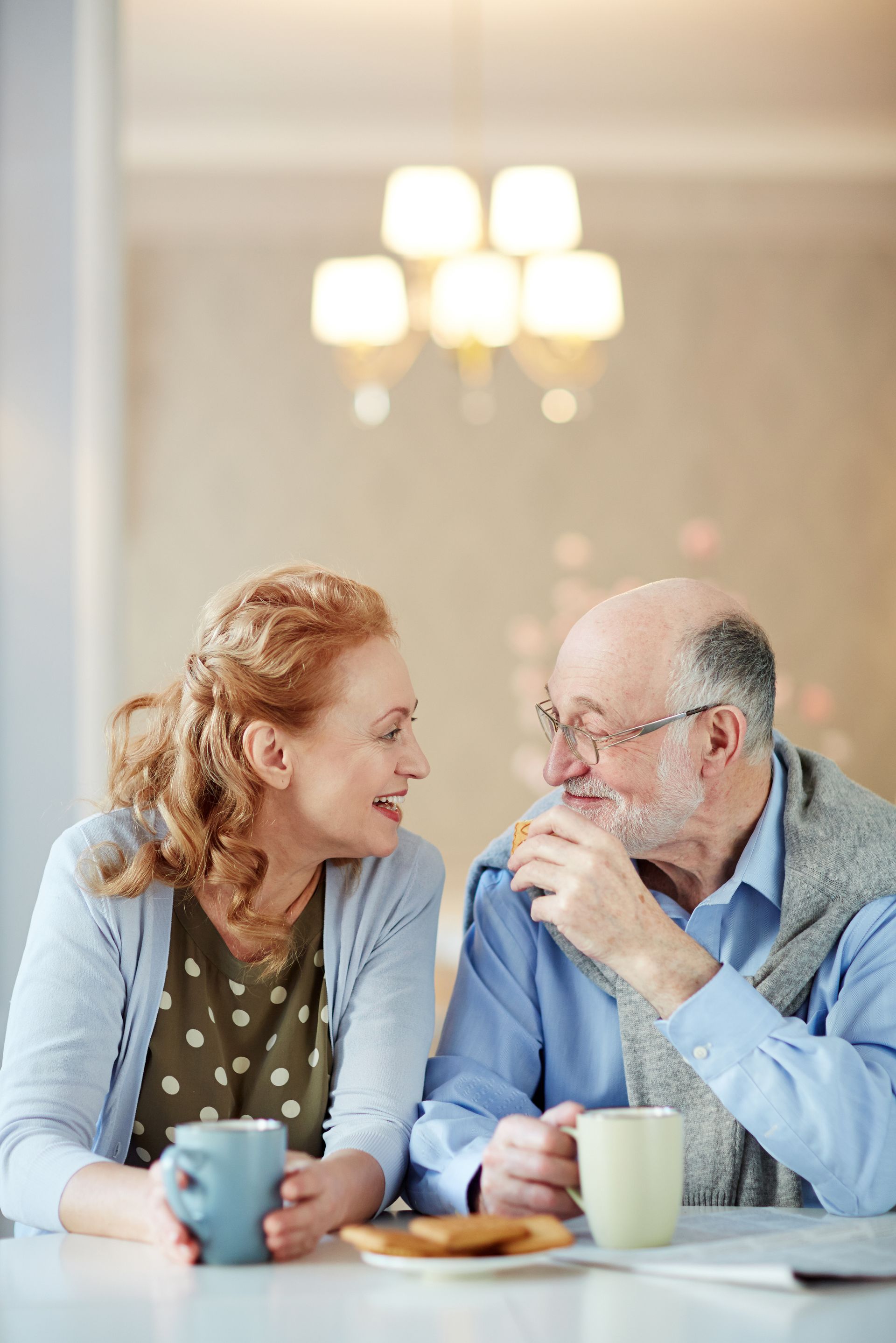 Elderly couple smiles and chats over coffee at a table indoors.