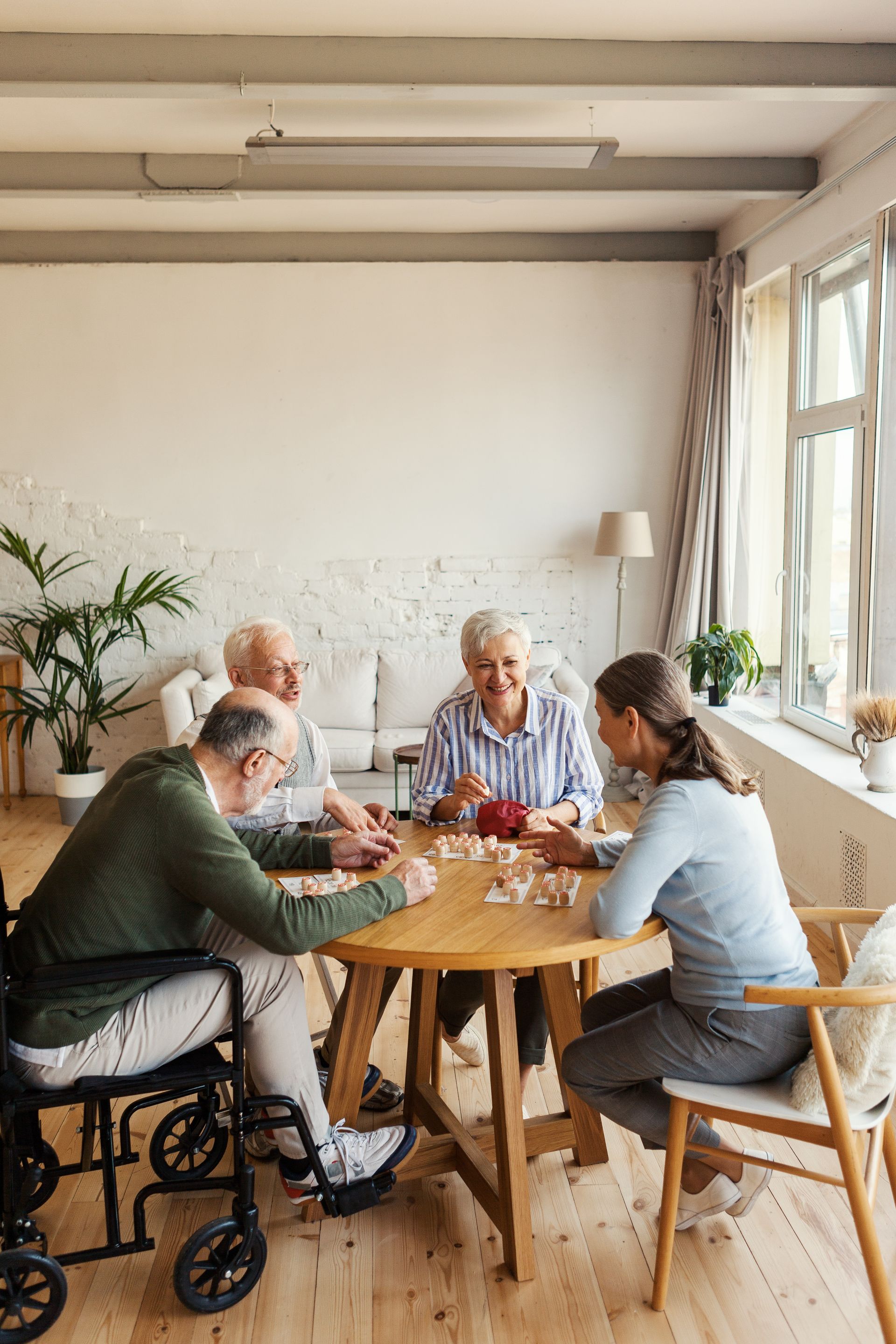 Group of seniors and a caregiver playing a game at a round table in a bright room with a window.