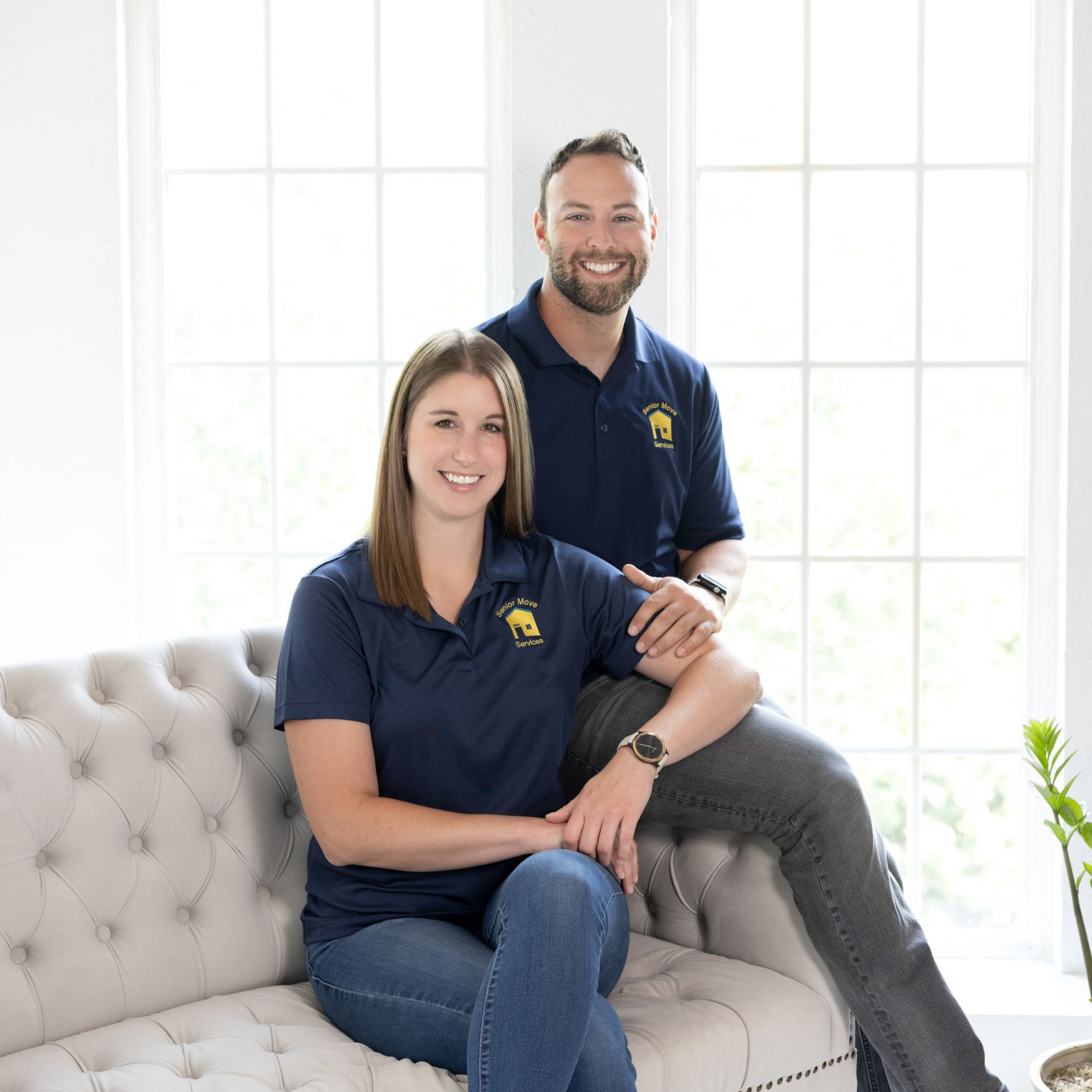 A man and woman in matching blue shirts sit on a couch in front of a window.