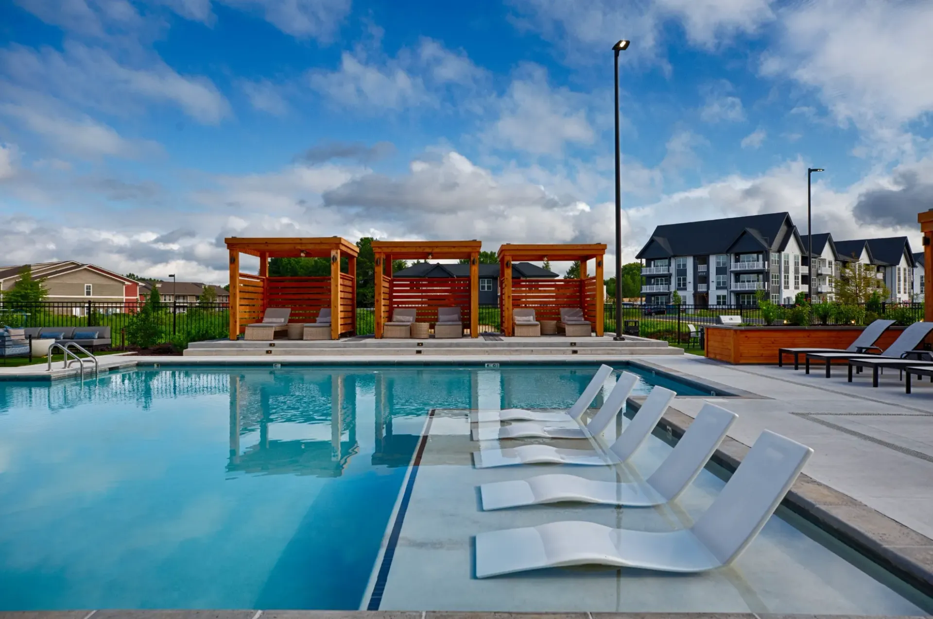 Pool with white lounge chairs, cabanas, and buildings under a blue sky.
