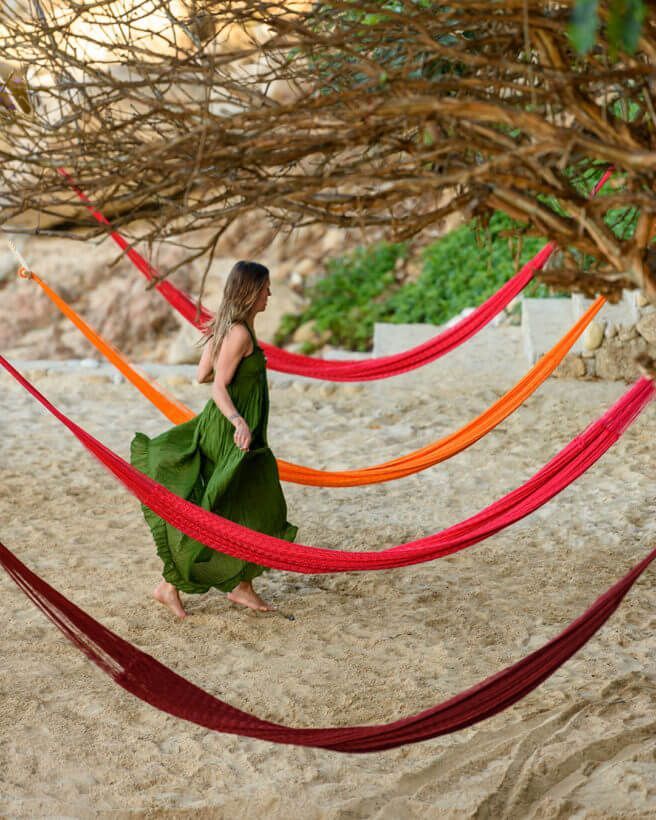 Woman in a green dress standing among colorful hammocks on a sandy beach.