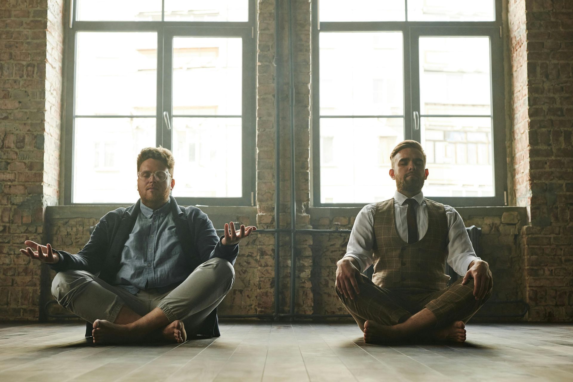 Two men meditating, cross-legged, in front of windows. One wears a blazer, the other a vest and tie. Brick wall setting.