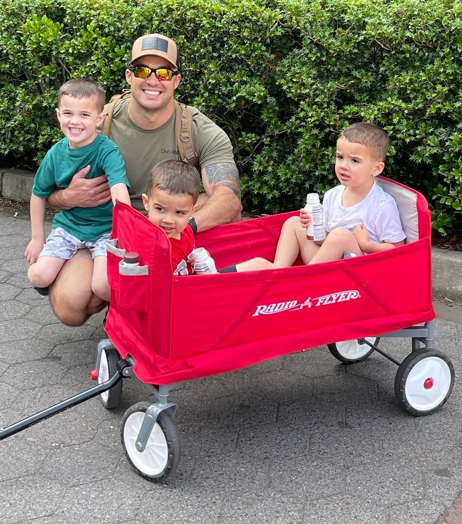 A man is kneeling next to a red wagon with two children in it.