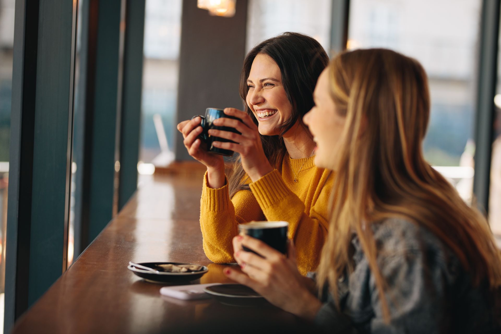 Two women laughing and holding coffee mugs at a sunlit cafe window. 