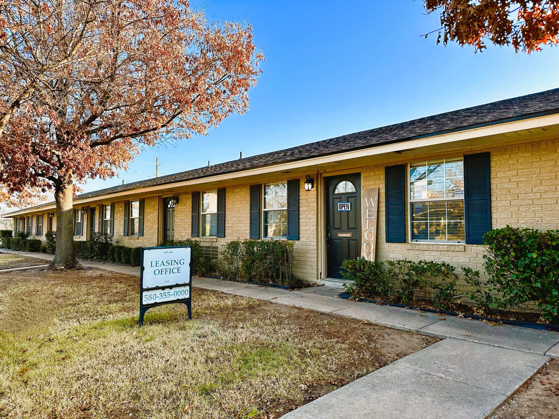 Apartment building with tan brick, blue shutters, and a sign on the lawn under a tree.
