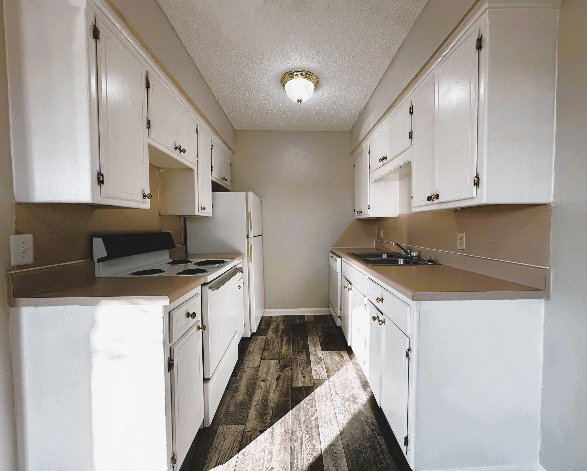 A narrow kitchen with white cabinets, appliances, and beige countertops. Dark wood-look flooring.