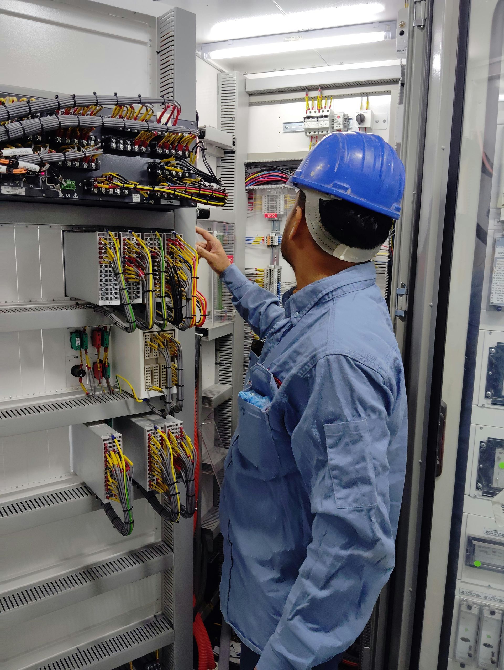 Technician in blue hard hat inspecting wiring inside an open electrical control panel.
