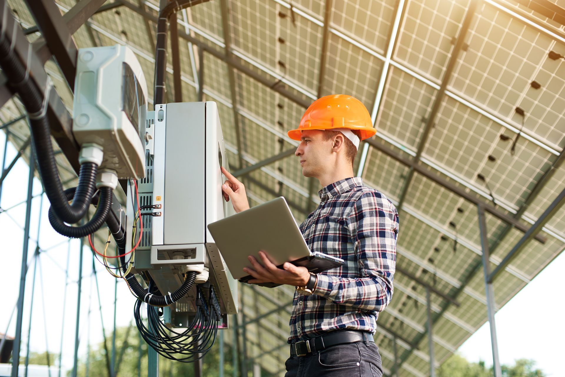 Technician in orange hard hat inspecting electrical equipment and taking notes in an industrial setting
