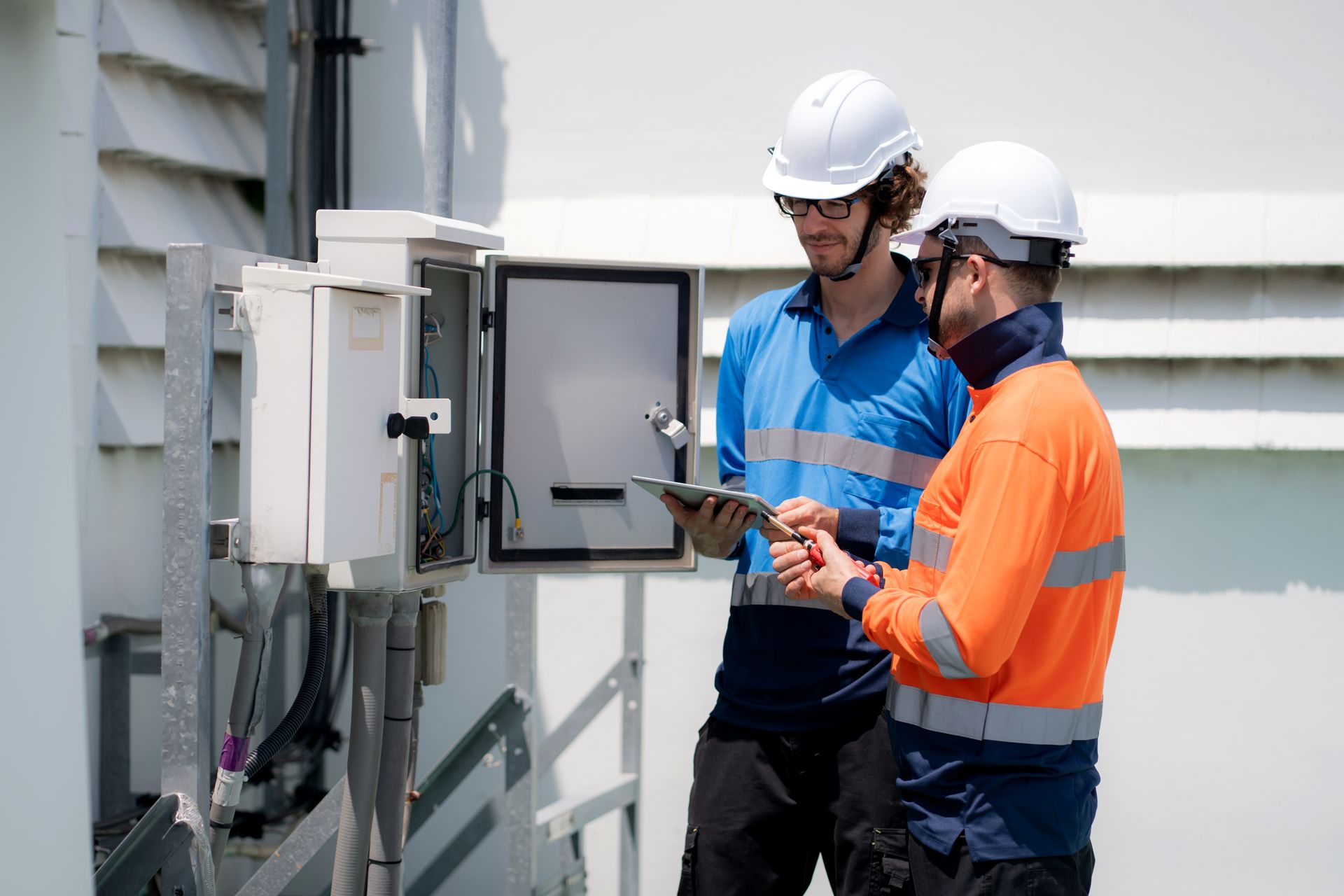 Two workers in hard hats inspect an open electrical control box outside a building.