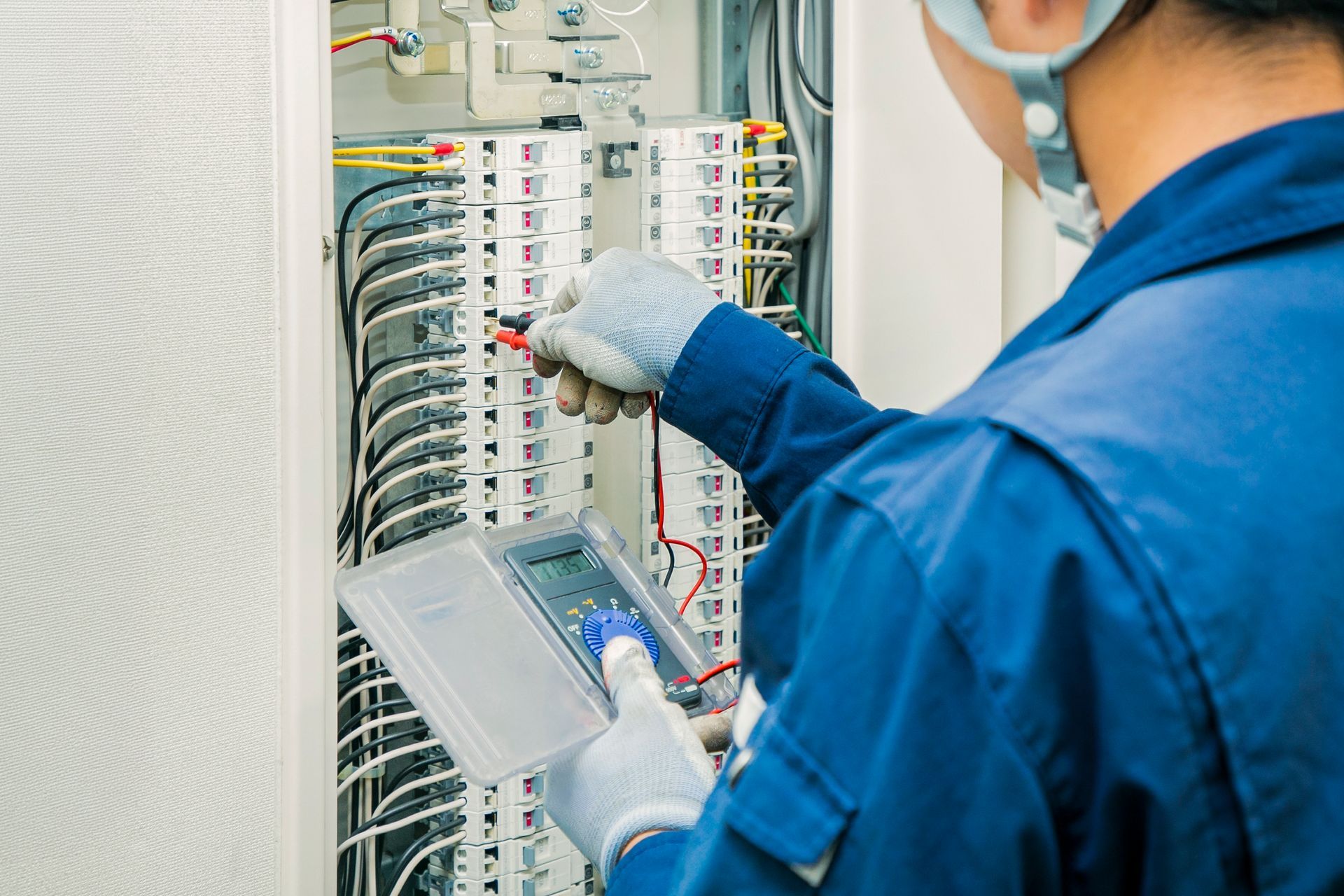 Technician in blue coveralls inspecting an electrical control panel with a handheld device