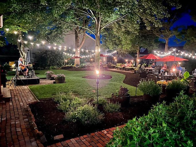 A live musician performs on a stage in an outdoor garden at dusk, illuminated by string lights and red patio umbrellas.