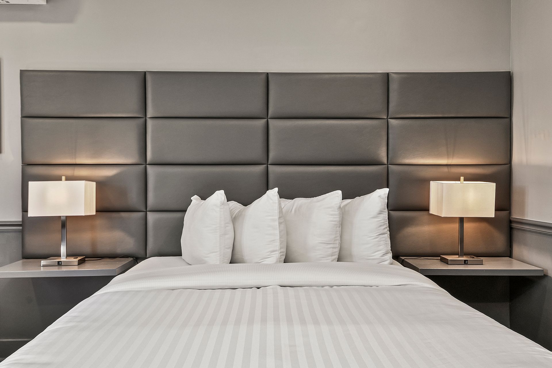 A bed with white bedding, pillows, and a large, gray, tufted leather headboard with two bedside lamps.