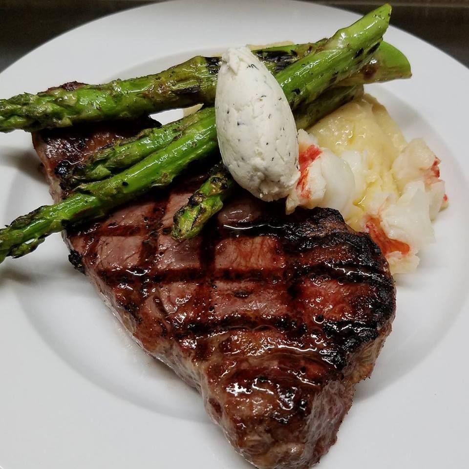 A grilled steak topped with a dollop of herb butter, accompanied by roasted asparagus and a side of lobster.