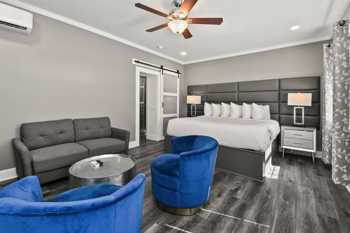 A modern hotel room with a large bed, a dark grey sofa, two blue velvet chairs, and a round metallic coffee table.